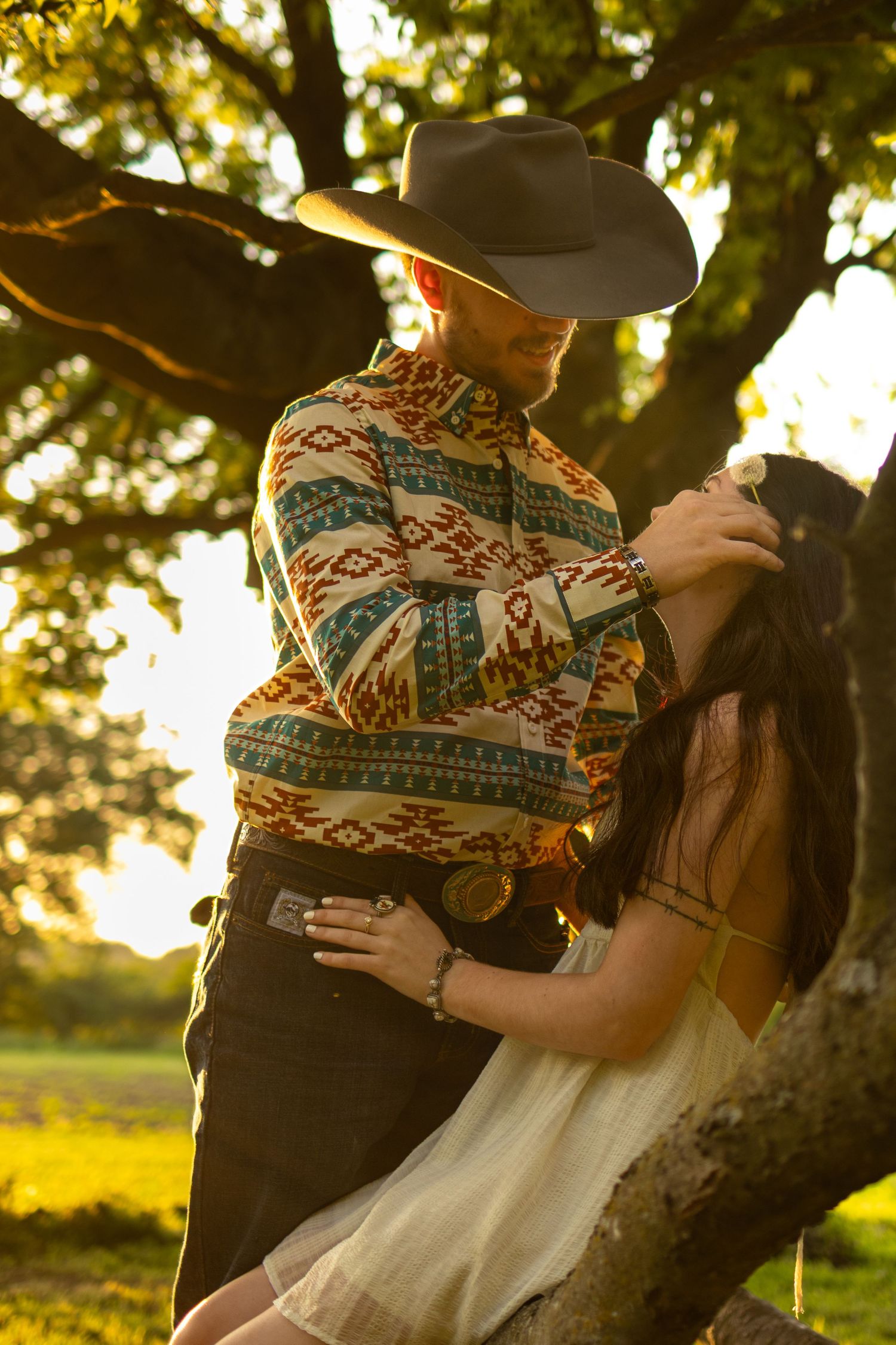 Couple embracing under a tree at golden hour, man in aztec print cowboy shirt and black hat, woman in white dress.