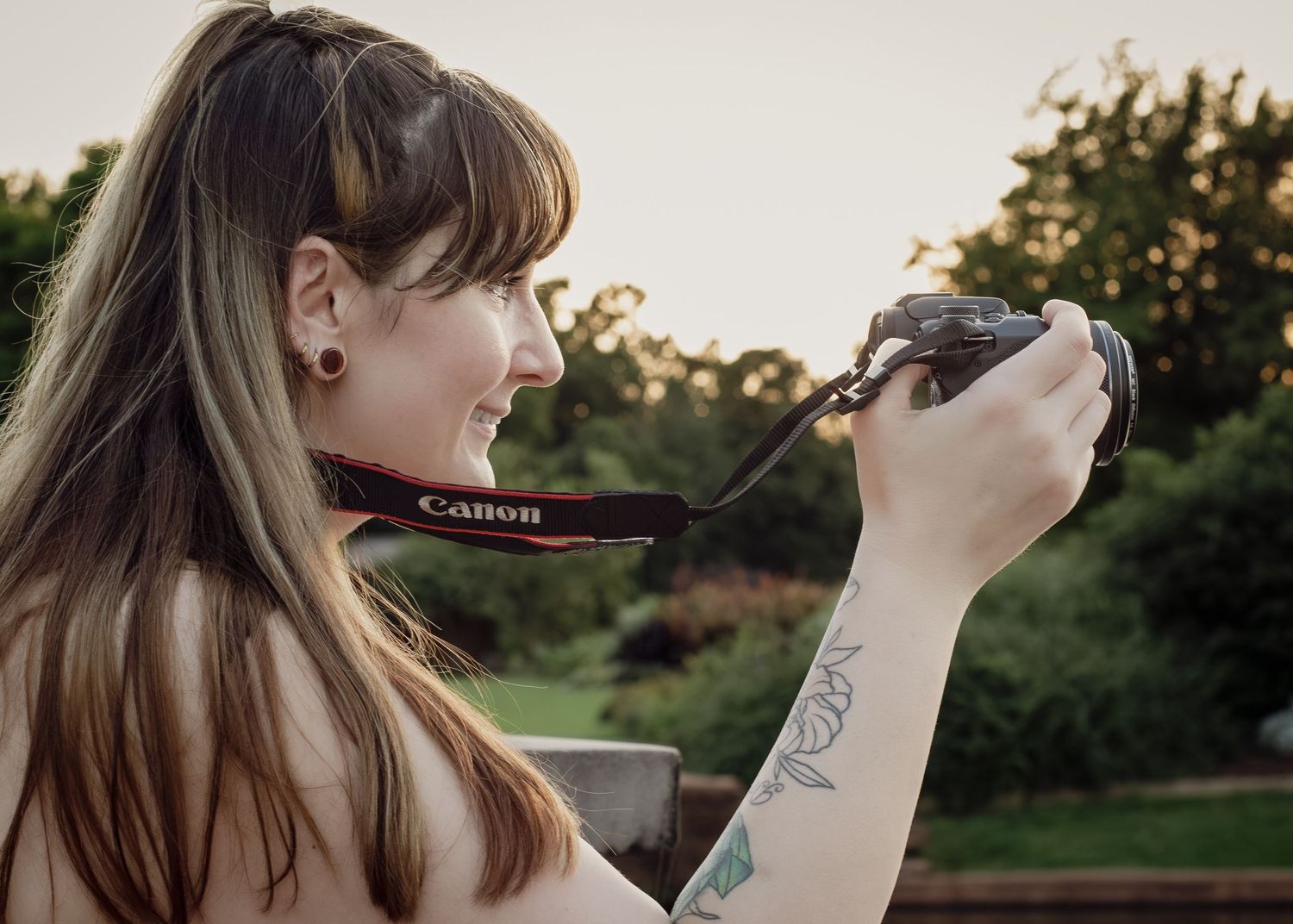 Smiling woman with tattoos holding a Canon camera outdoors, with green trees and warm sunset light in the background.