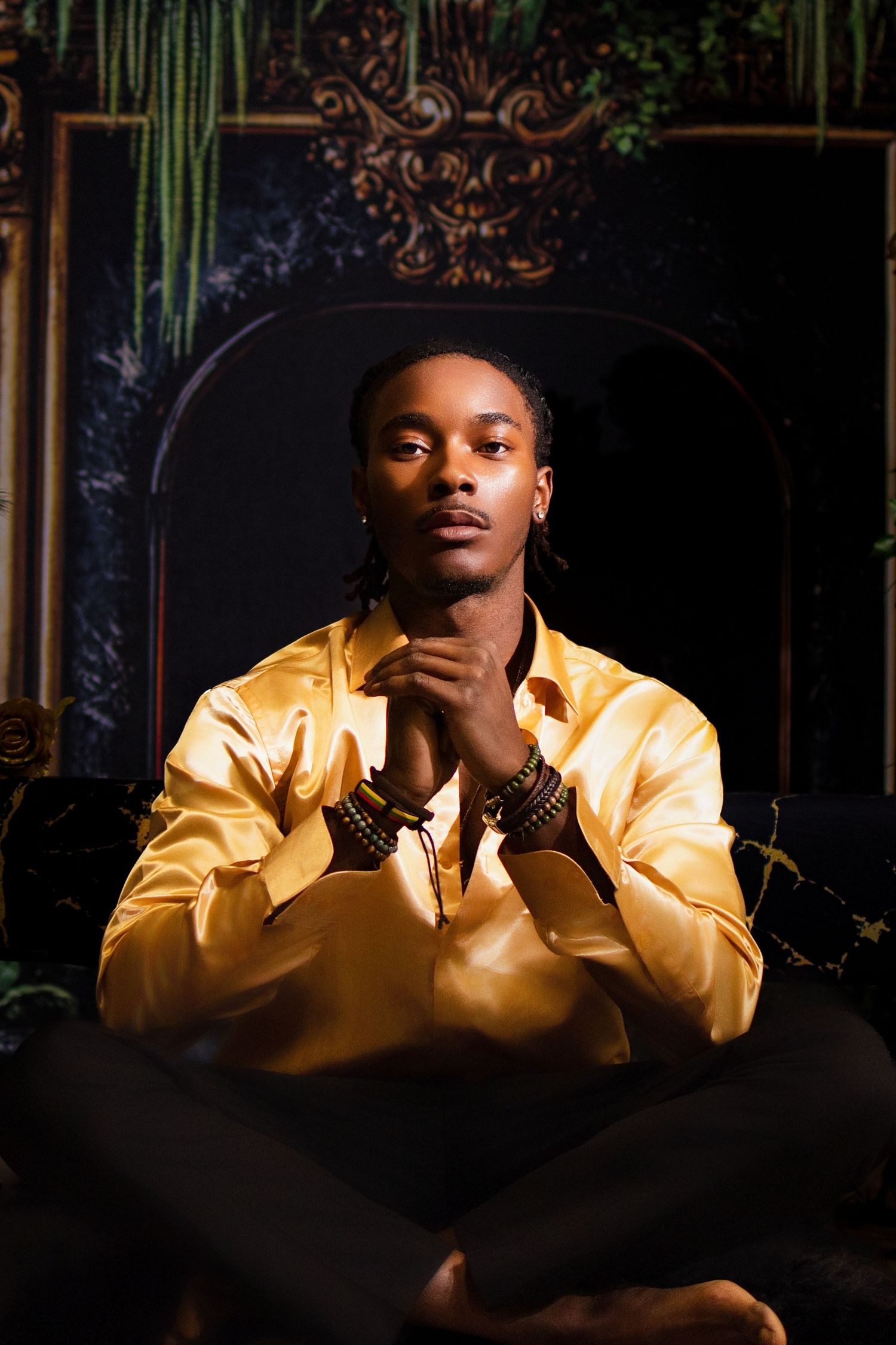 Young man in golden satin shirt sitting cross-legged, hands clasped, posed before ornate dark archway backdrop.