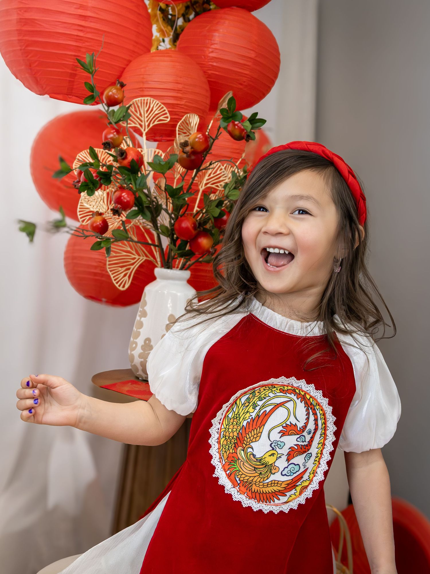 Young girl in red dragon dress laughs joyfully amid Chinese New Year red lanterns and berry decorations.