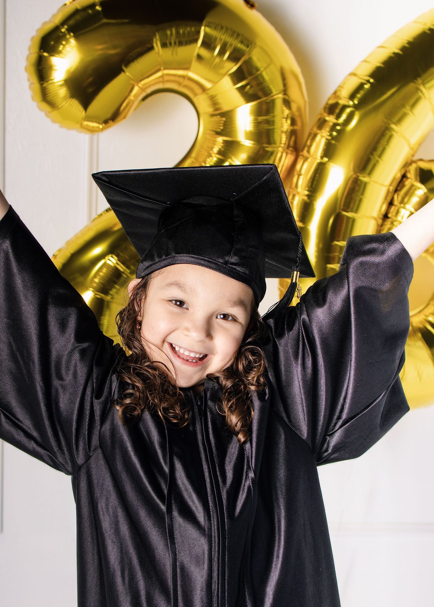 Excited young girl in black graduation cap and gown celebrating with arms raised near gold balloons.