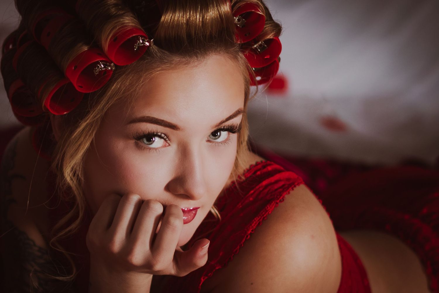 Young blonde woman with red hair rollers wearing red velvet, posing elegantly with hand near face.
