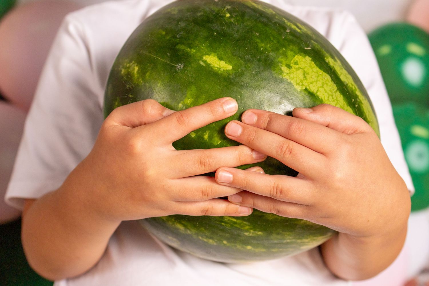 Child holding a large green watermelon with both hands, wearing a white shirt, with green balloons in the background.