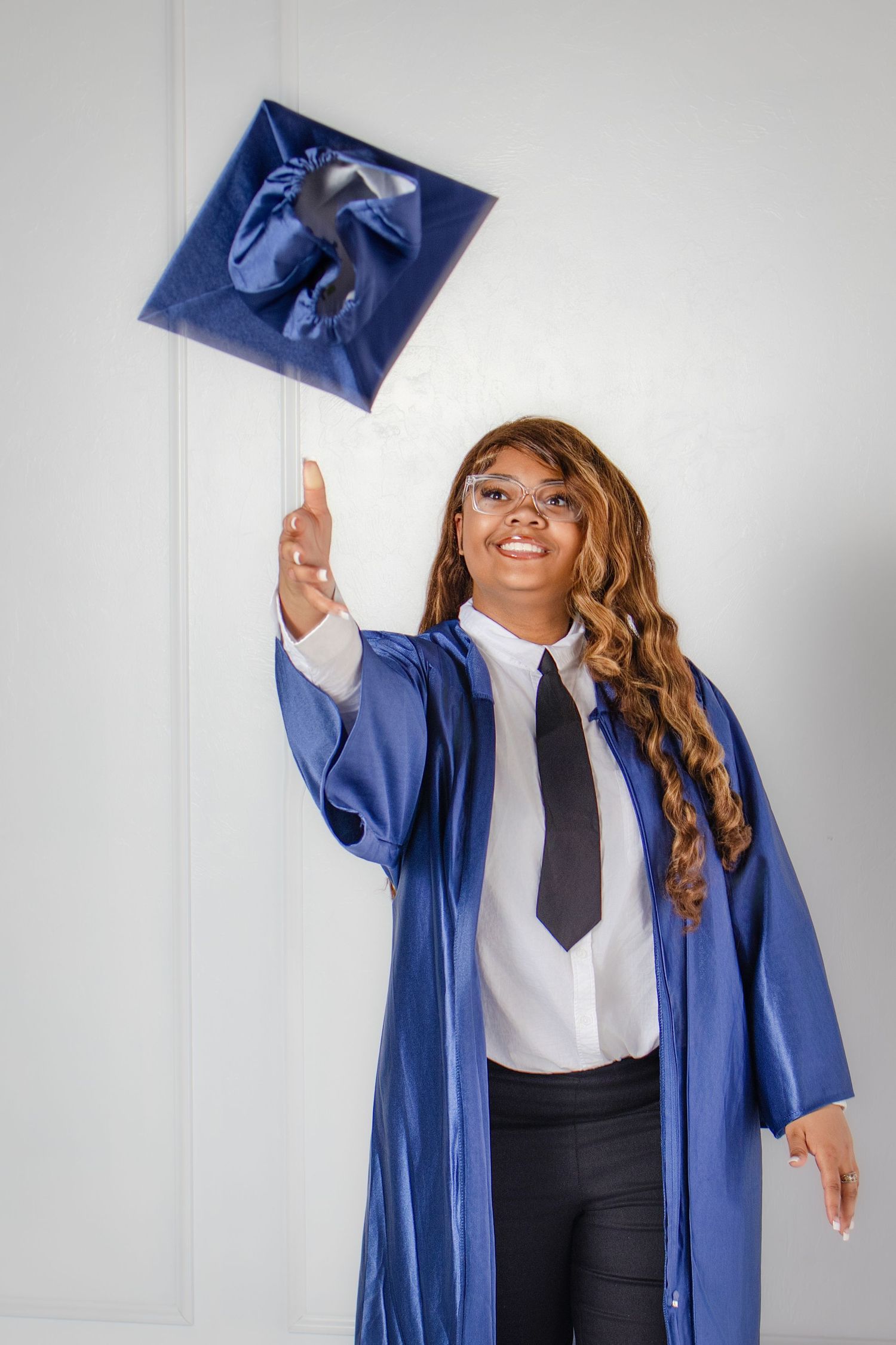 Smiling graduate in blue cap and gown tossing her cap in the air, celebrating her academic achievement.