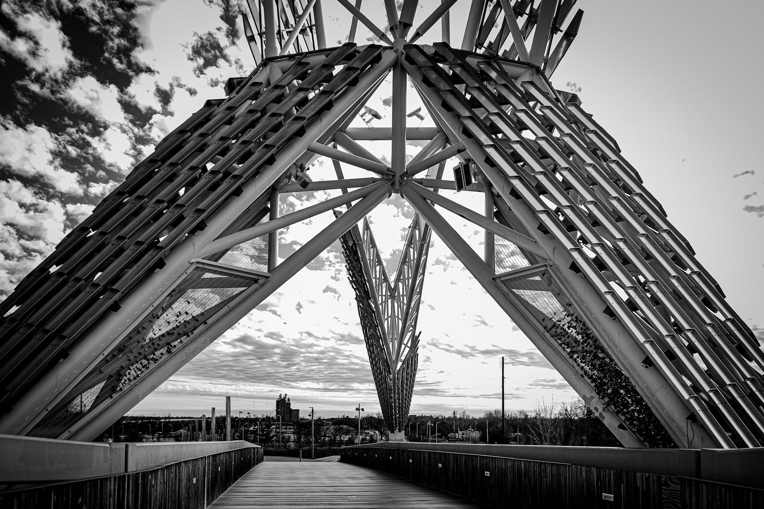 Black and white photo of a geometric steel bridge structure with dramatic sky and cityscape in background.