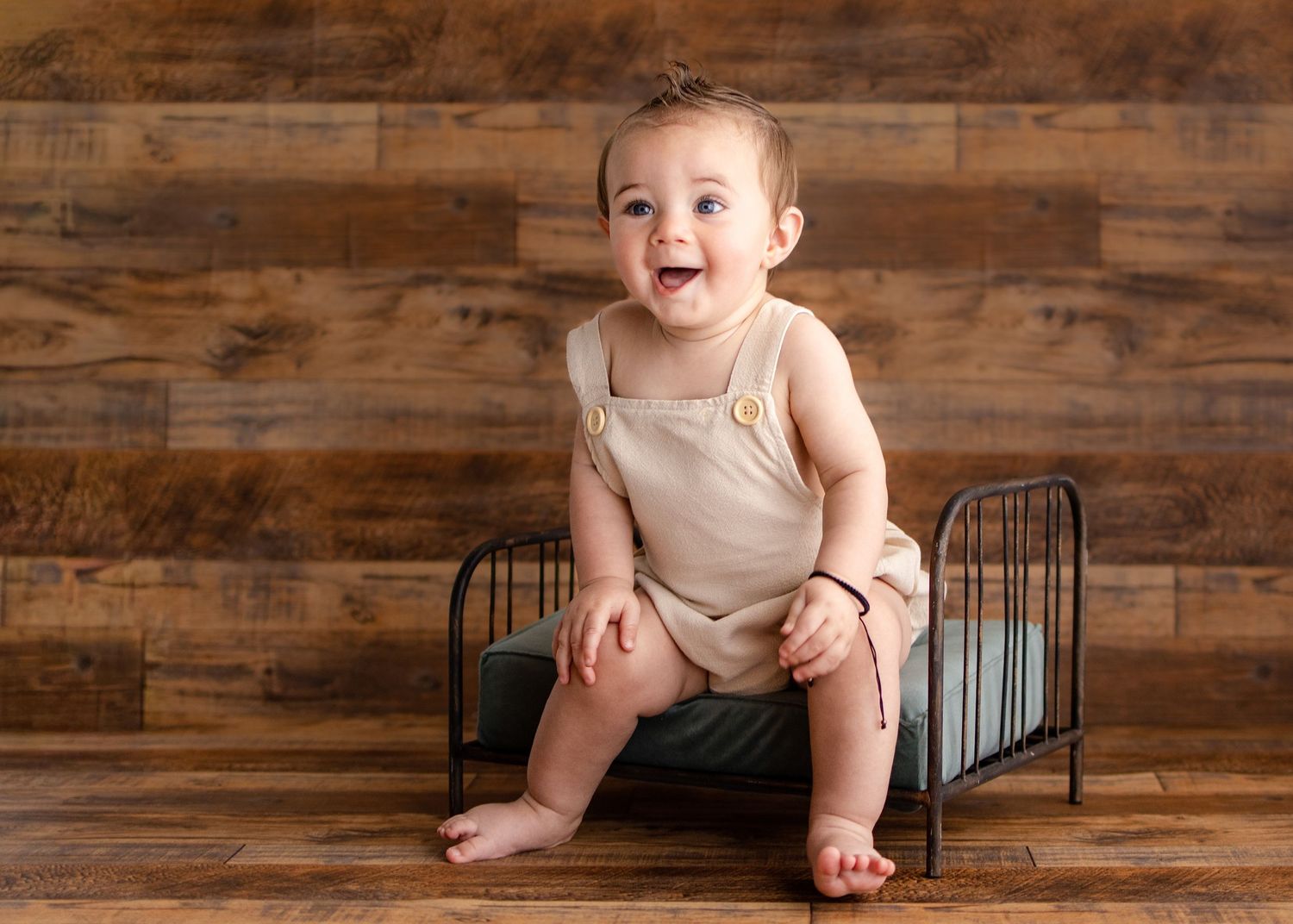 Smiling baby boy in beige romper sitting on a small metal bed frame against a rustic wood plank backdrop.