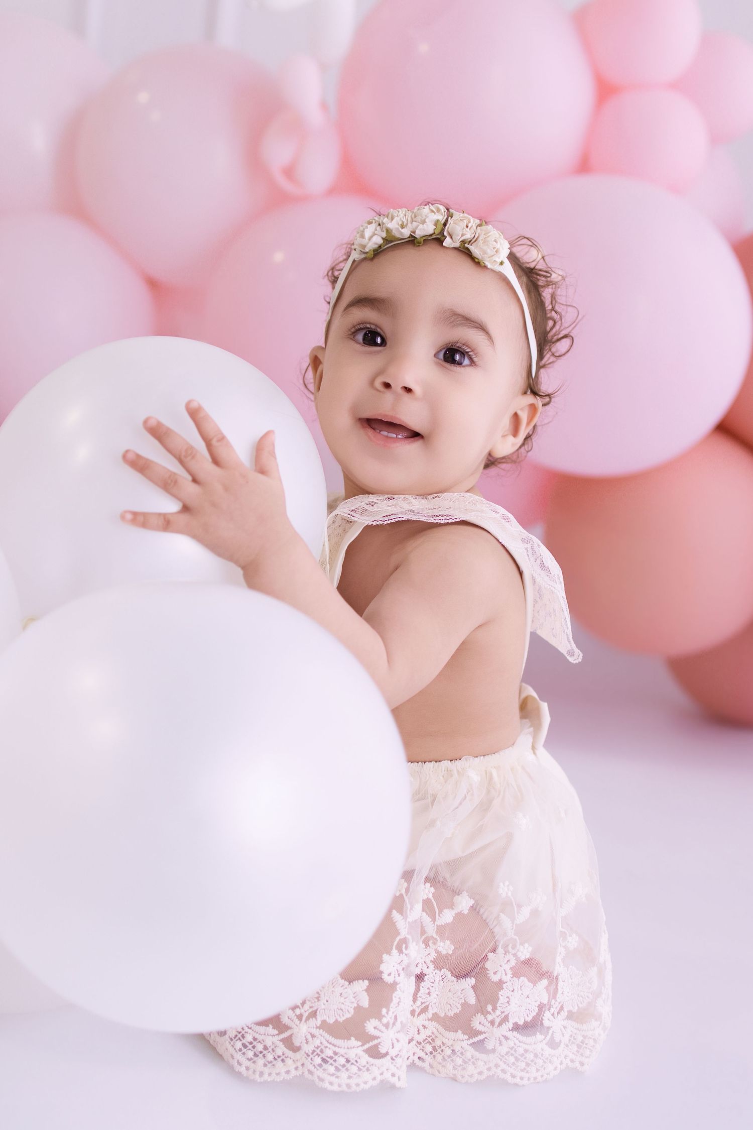 Adorable baby girl in lace outfit and floral headband playing with white balloons at a pink balloon party.