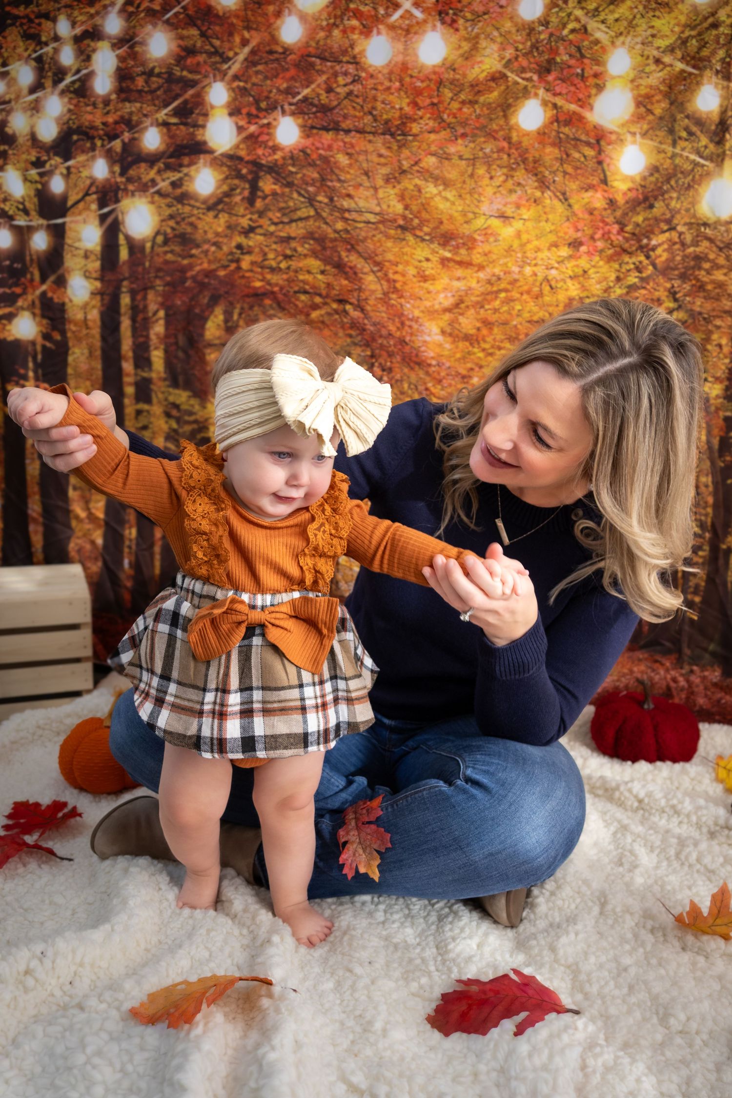 A mother helps her baby girl stand during a fall-themed photo session with autumn leaves and warm bokeh lights.