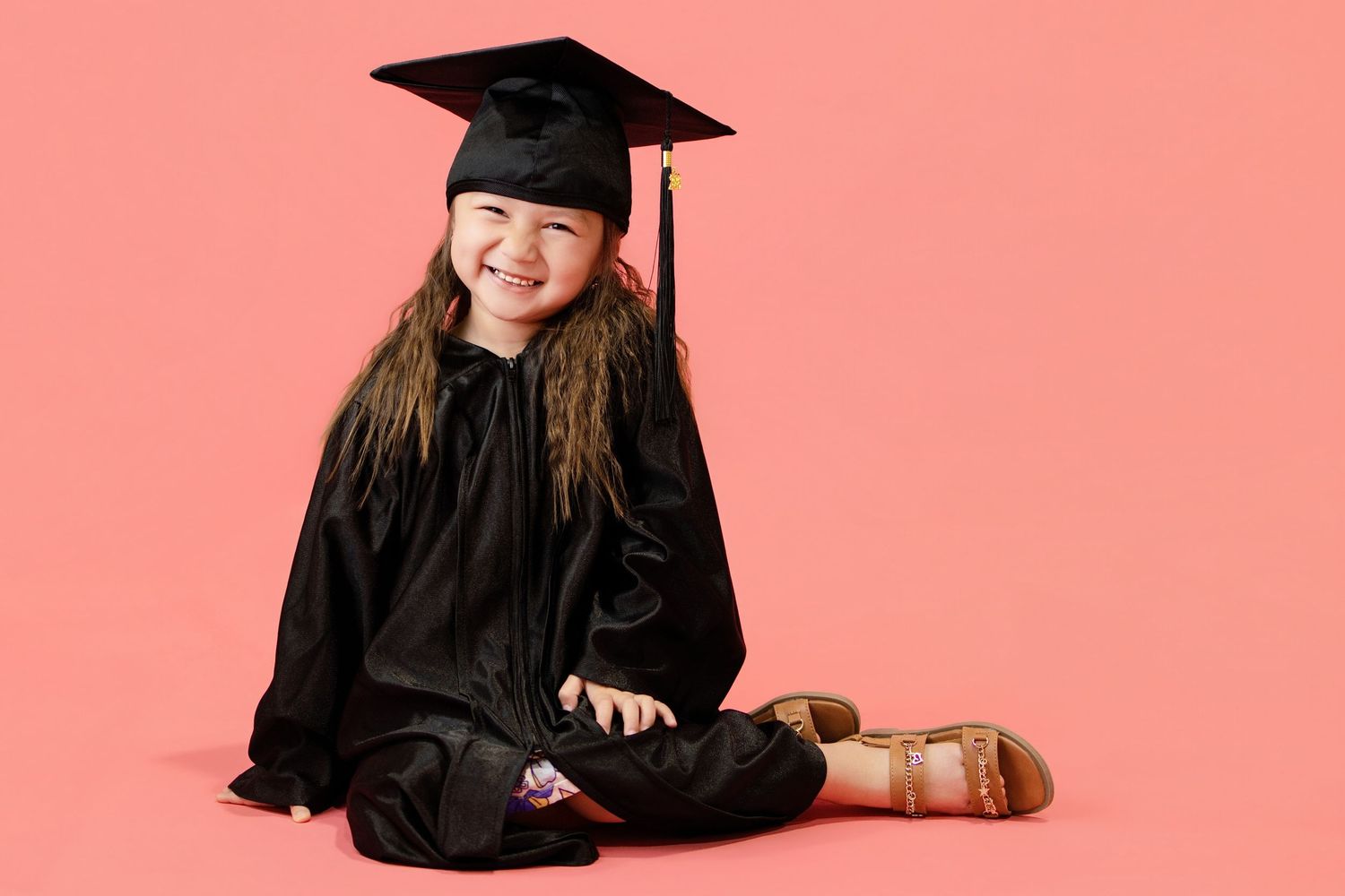 Happy young girl sitting cross-legged in a black graduation gown and cap on a pink background.