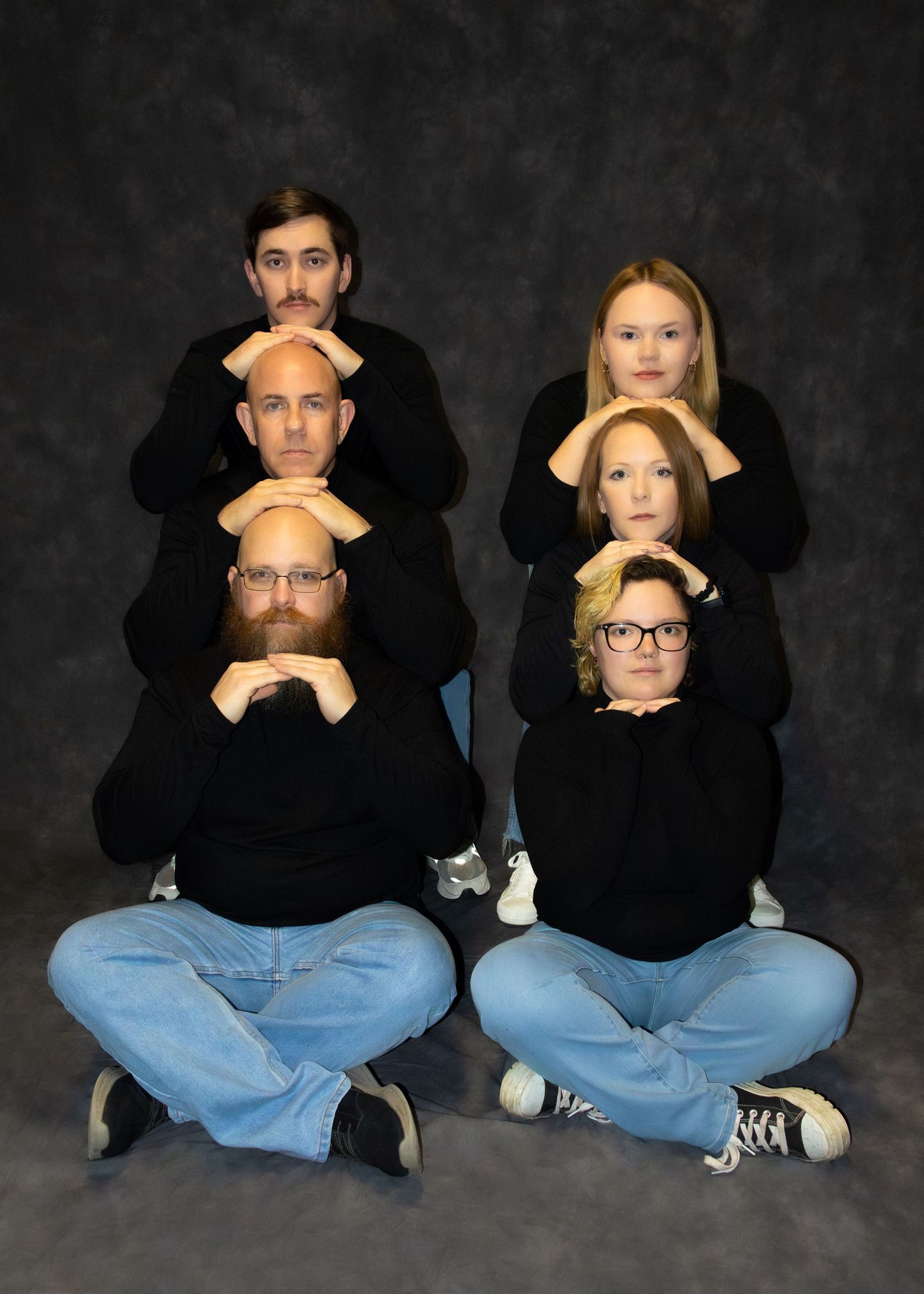 Six people in black sweaters pose creatively stacked in a humorous group portrait against a gray studio background.