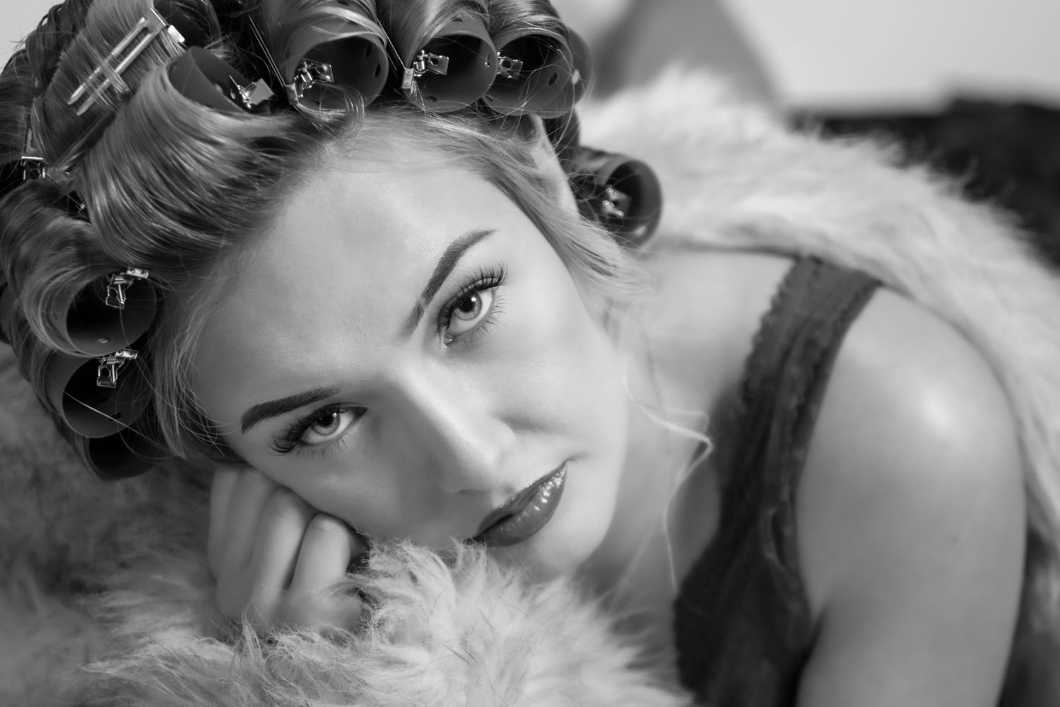 Black and white portrait of a woman with hair curlers resting her chin on her hand on a fur surface.