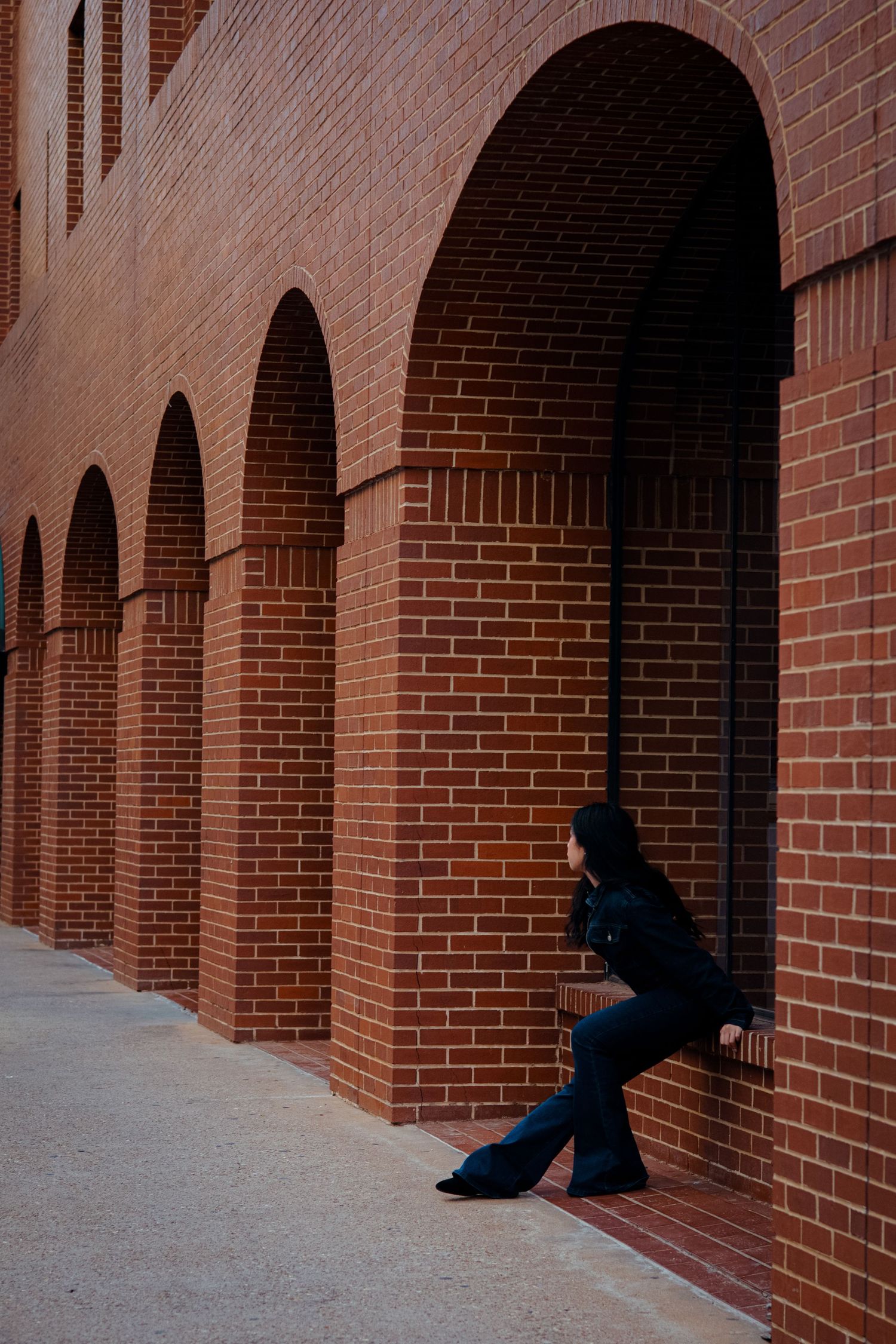 Woman in dark jeans leaning against red brick building with arched alcoves.
