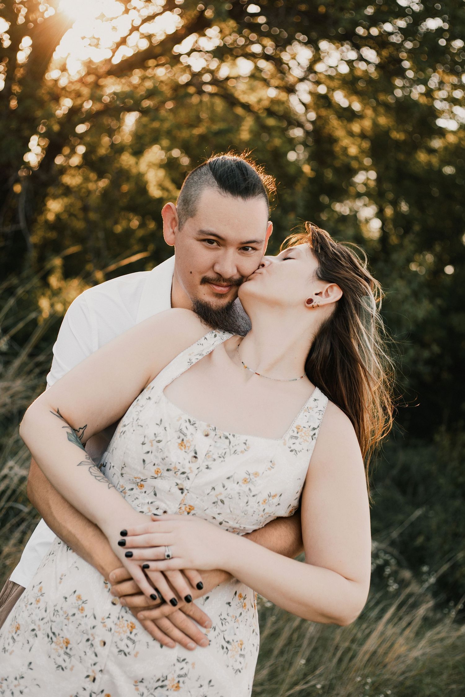 Couple embracing at golden hour, woman in floral dress kissing man's cheek, surrounded by lush green trees.
