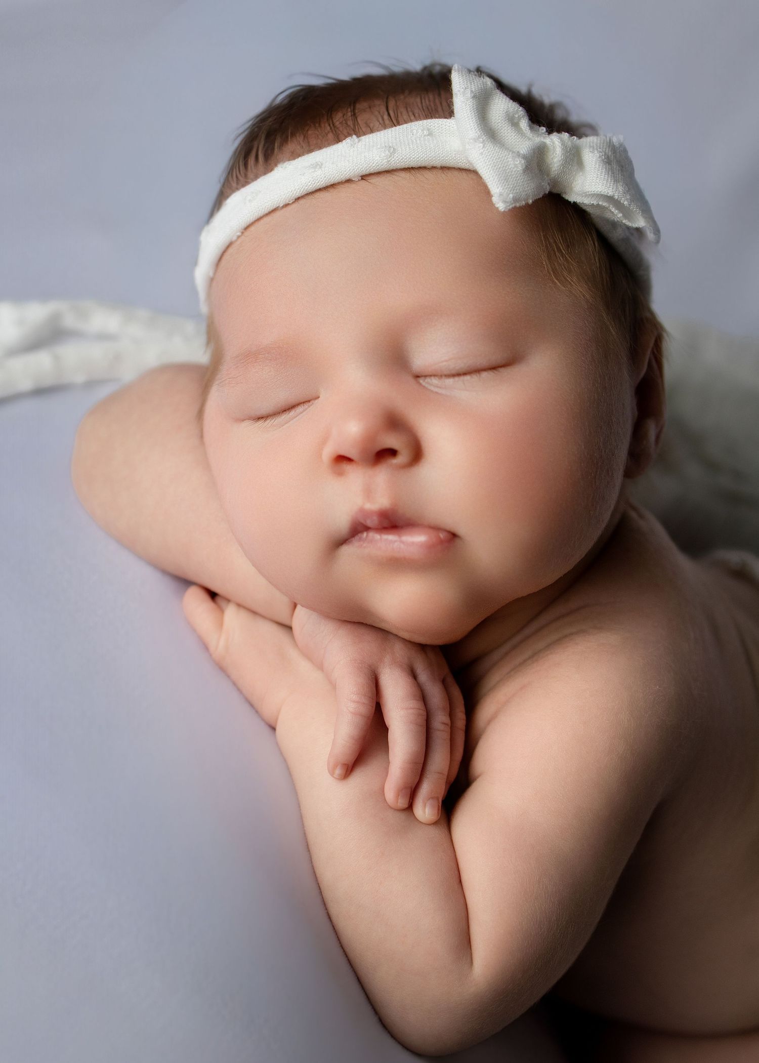 Peaceful sleeping newborn baby girl wearing a white bow headband, posed with hands under chin on soft gray background.