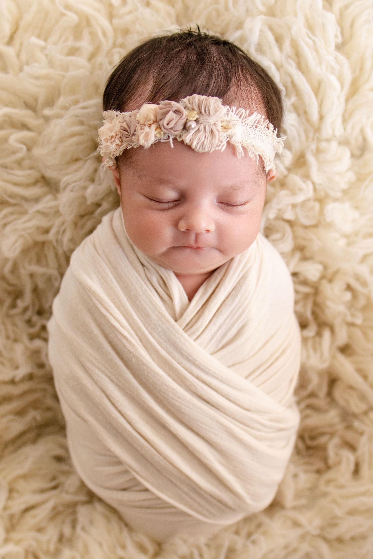 Sleeping newborn baby swaddled in cream wrap with floral headband on fluffy white backdrop.
