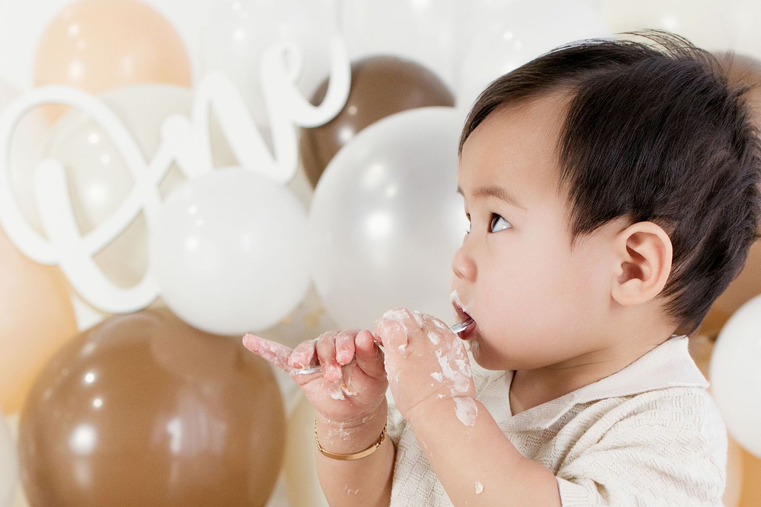 A baby celebrates their first birthday smashing cake with balloons and a cursive 