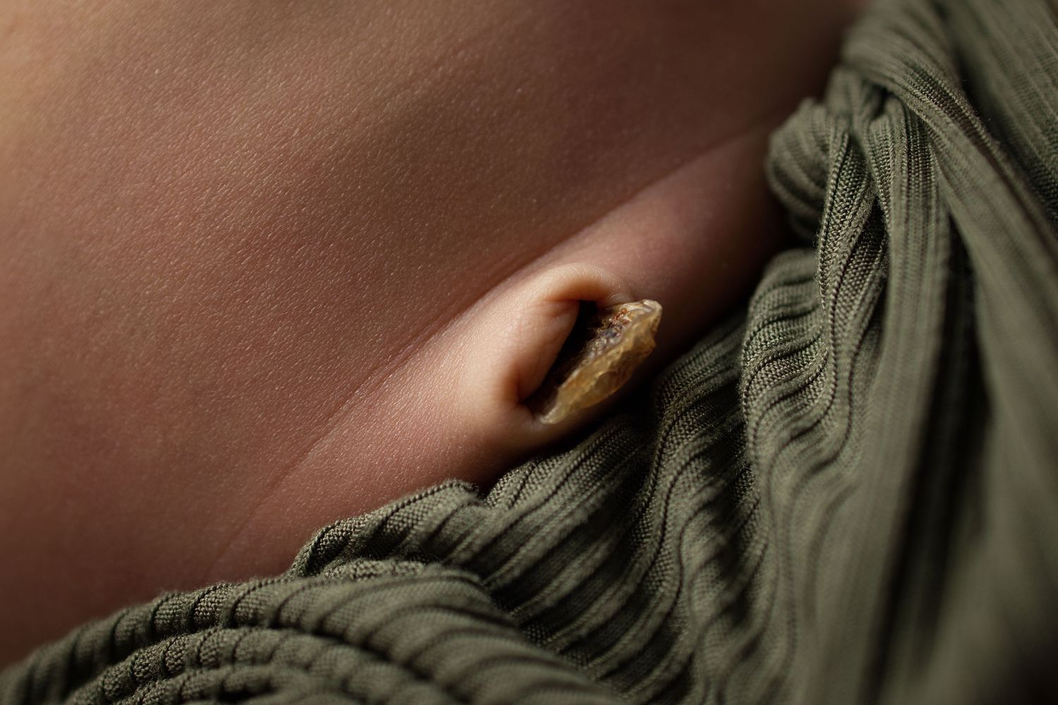Close-up of a human navel with a small brown belly button ring against bare skin and olive green fabric.