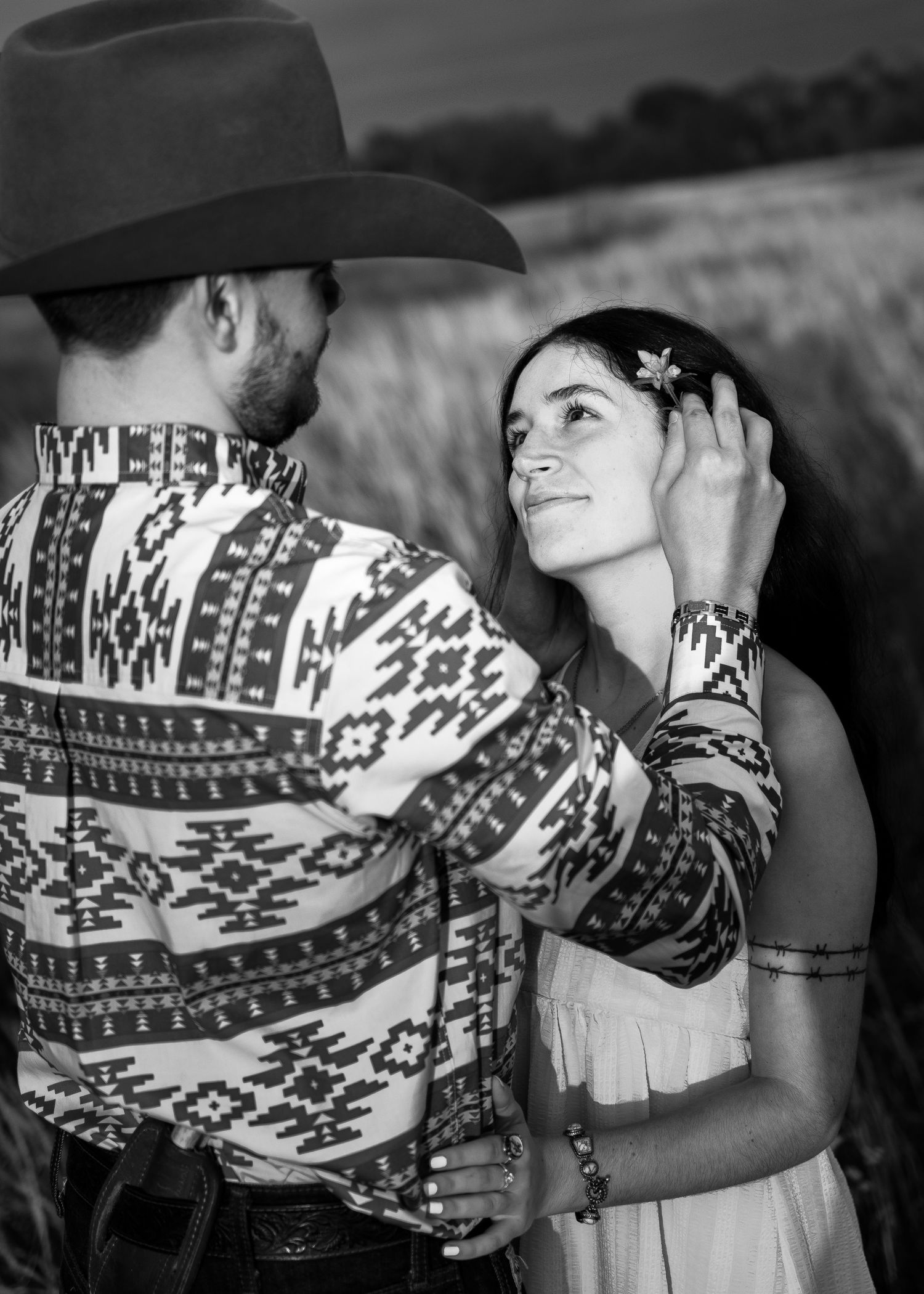 Black and white photo of a couple in western attire sharing a tender moment in a field, man in cowboy hat touching womans hair.