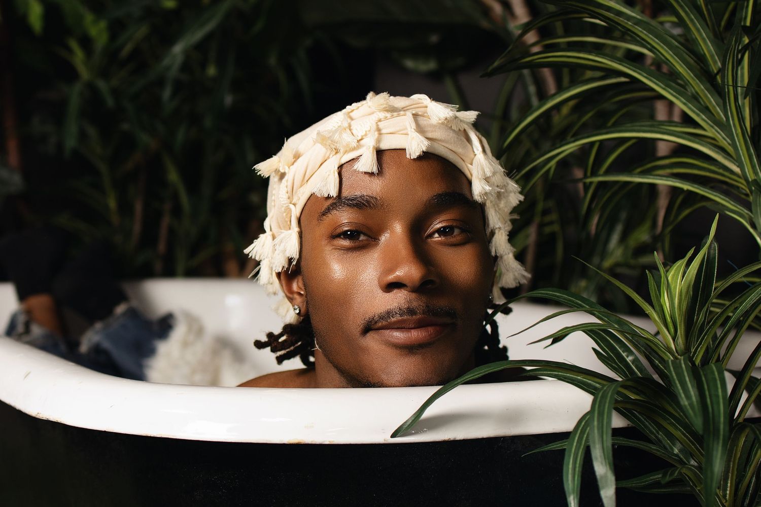 Young man relaxing in a bathtub wearing a shower cap, surrounded by lush tropical plants.