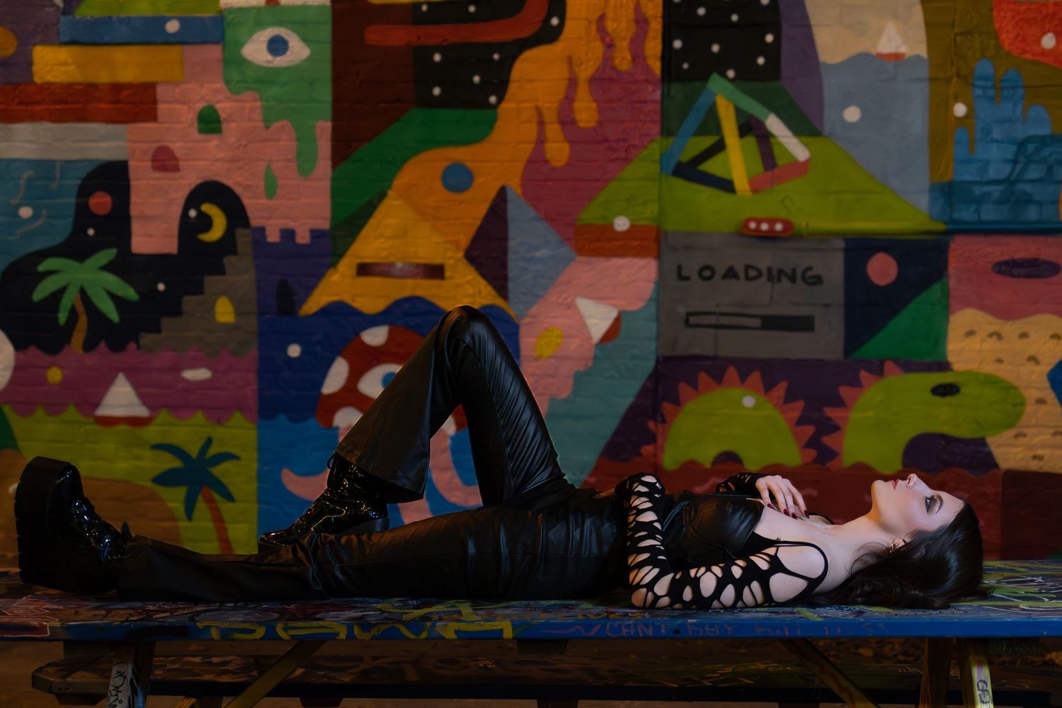 Woman in black leather outfit lying against a colorful graffiti mural wall.