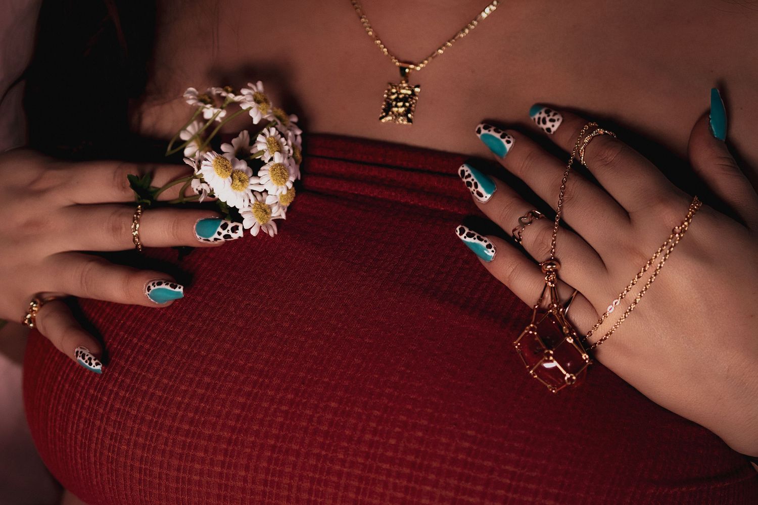 Close-up of hands with teal stiletto nails, gold rings, bracelets, and a floral ring, resting on a red ribbed top.