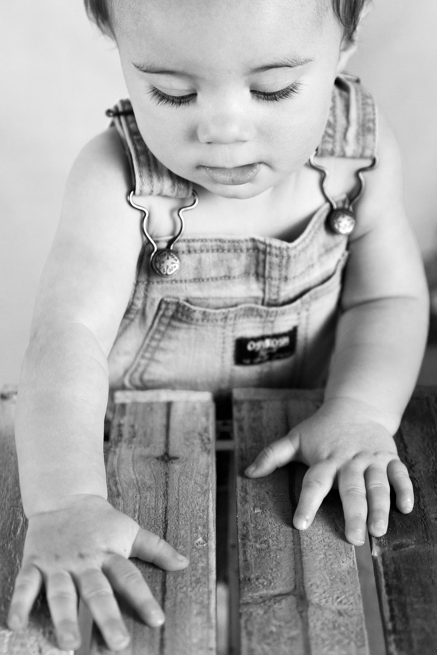 Black and white photo of a baby in OshKosh overalls leaning over a wooden surface.