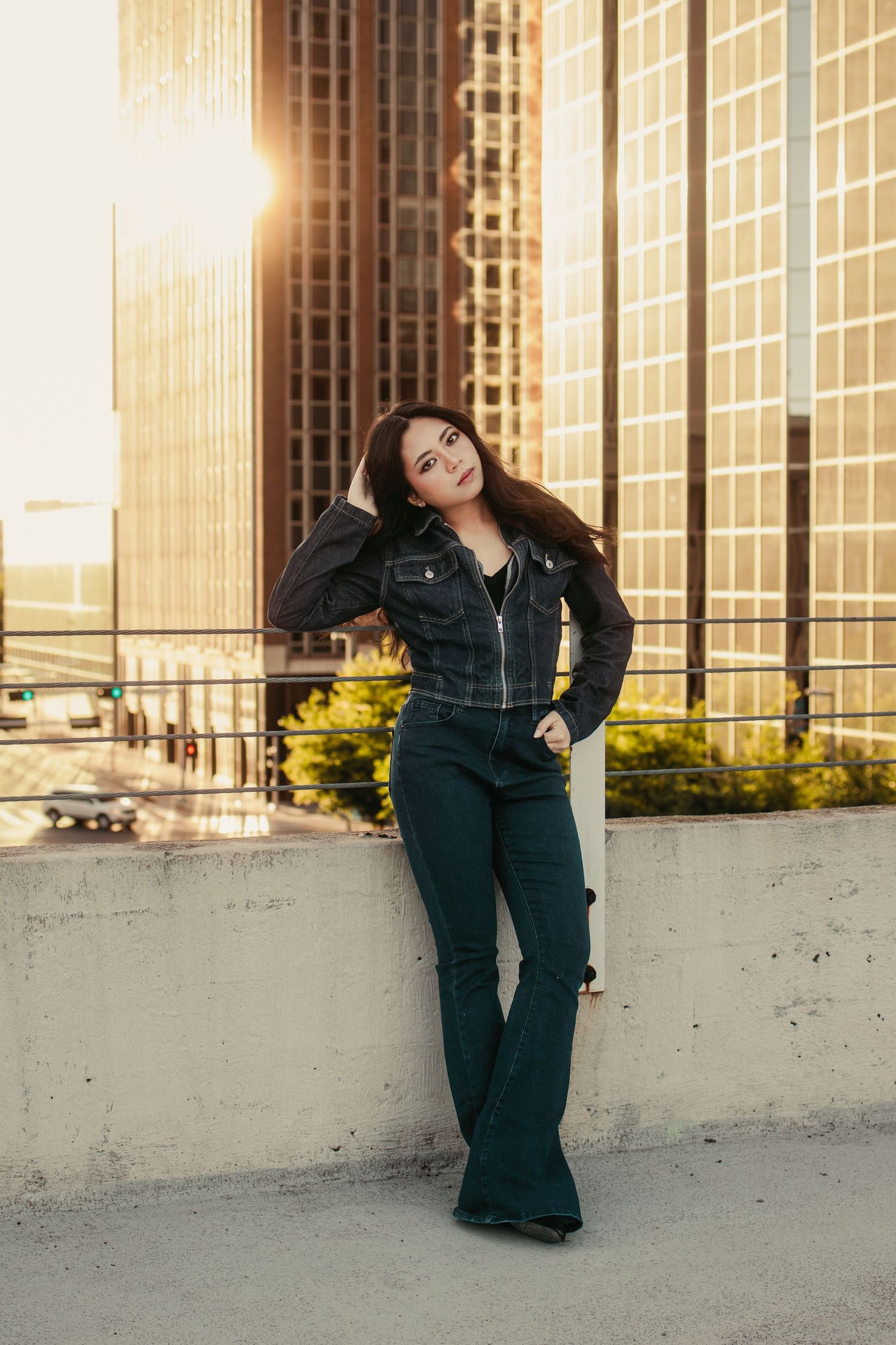 Young woman in dark denim outfit poses against rooftop wall with golden sunset and city skyscrapers behind her.