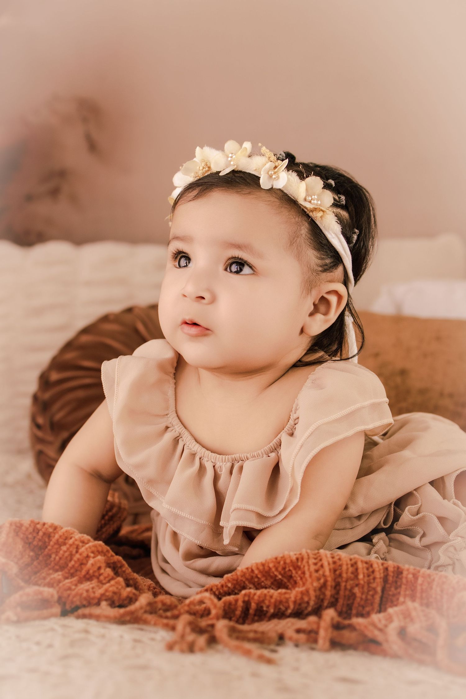 Adorable baby girl in beige ruffle romper and floral headband sitting in a wicker basket on a soft blanket.