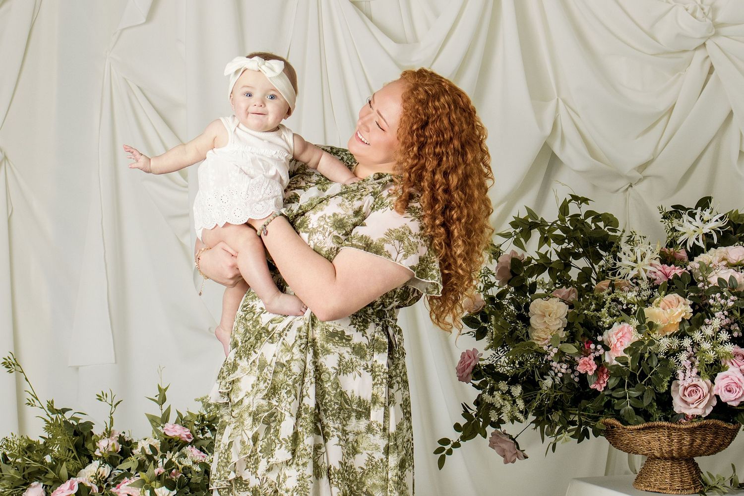 A curly redhead woman in a floral dress holds a smiling baby girl aloft, surrounded by lush pink and white floral arrangements.