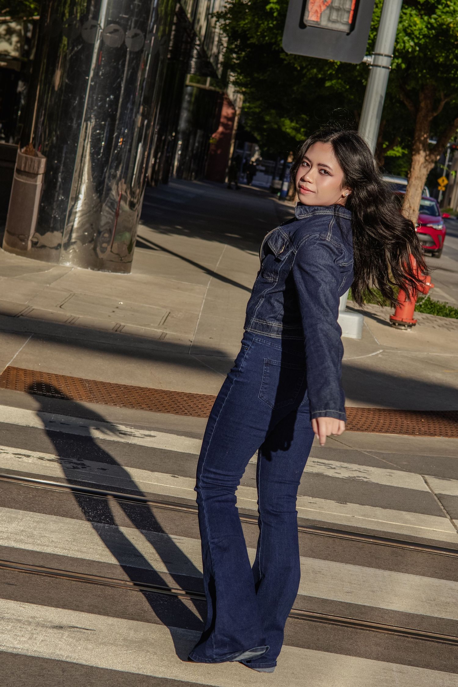 Stylish woman in dark denim jacket and flare jeans poses on city crosswalk in sunlight.