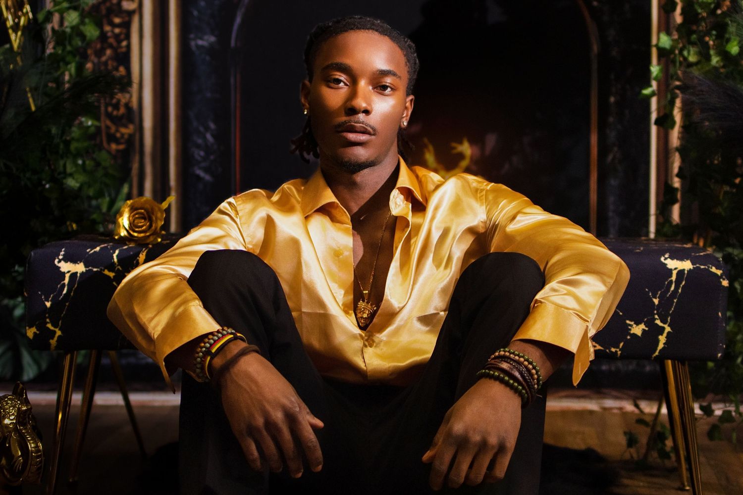 Young man in gold satin shirt seated on dark chair with lush green backdrop and ornate fireplace.
