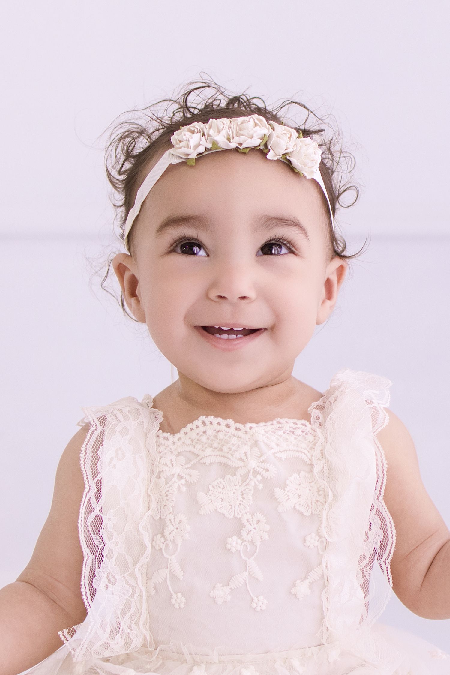 Smiling baby girl in white lace dress and floral headband portrait.