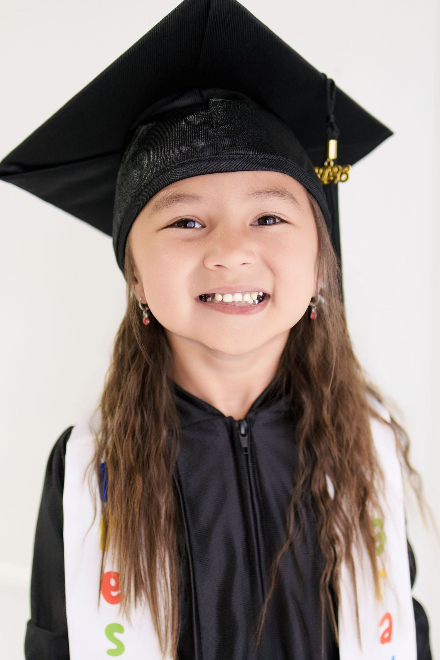 Happy young girl wearing black graduation cap and gown with colorful sash, smiling brightly.