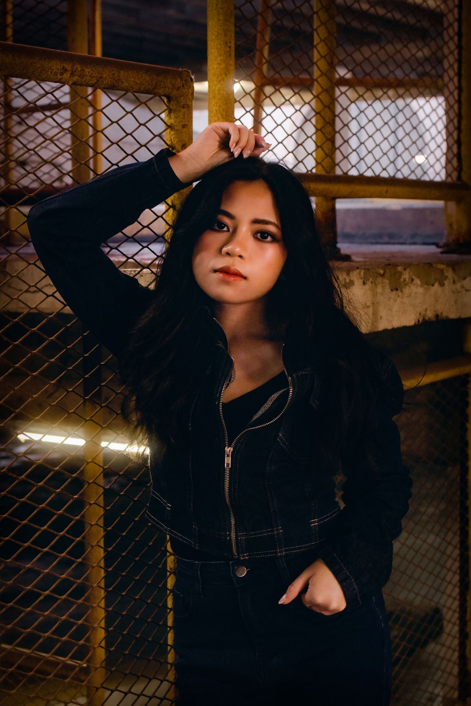 Young woman in black jacket poses against chain-link fence in dramatic urban night setting.