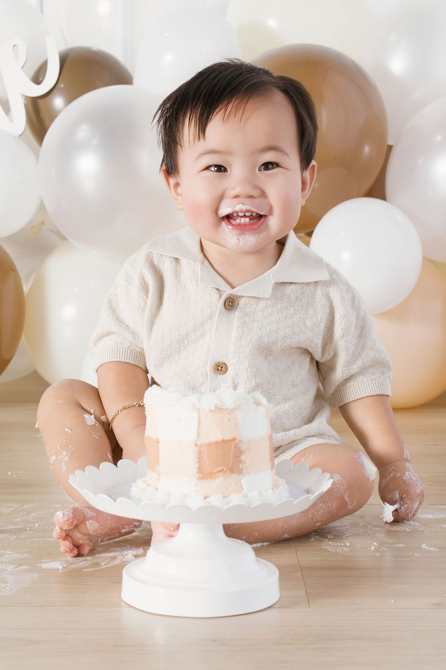 Smiling baby boy in beige outfit celebrating first birthday cake smash with white and gold balloons backdrop.