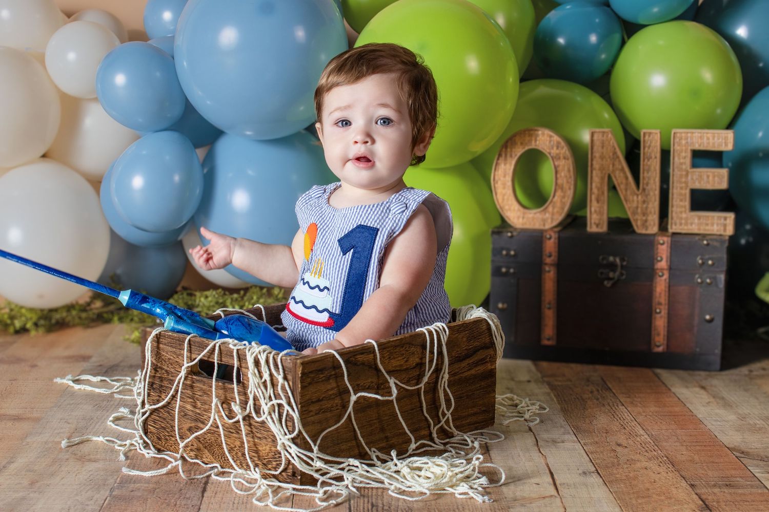 Baby boy sits in wooden box with fishing rod celebrating first birthday with ONE sign and balloons.