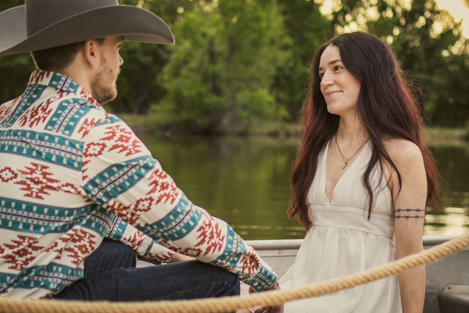 A couple shares a romantic moment on a boat, man in cowboy hat and patterned shirt, woman in white dress smiling.