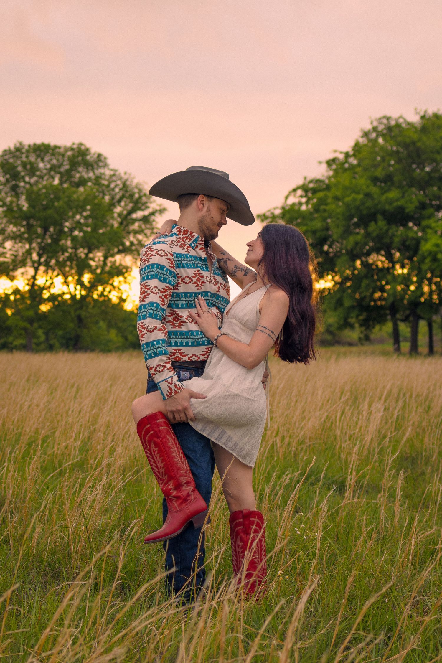 Couple embracing in tall grass field at sunset, man in cowboy hat and woman in red boots and white dress.