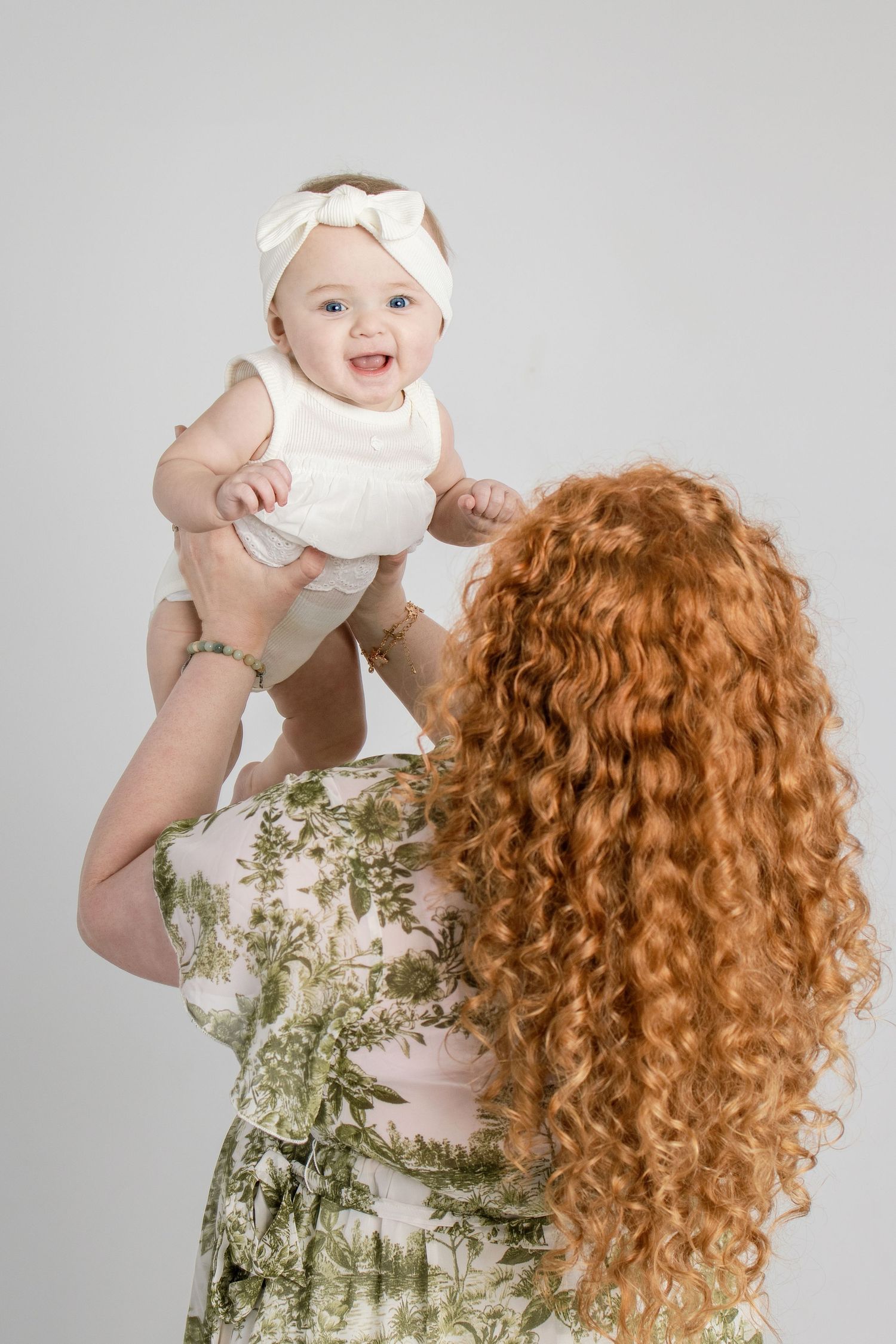 Redhead mother lifting smiling baby girl in white dress and bow headband against gray background.