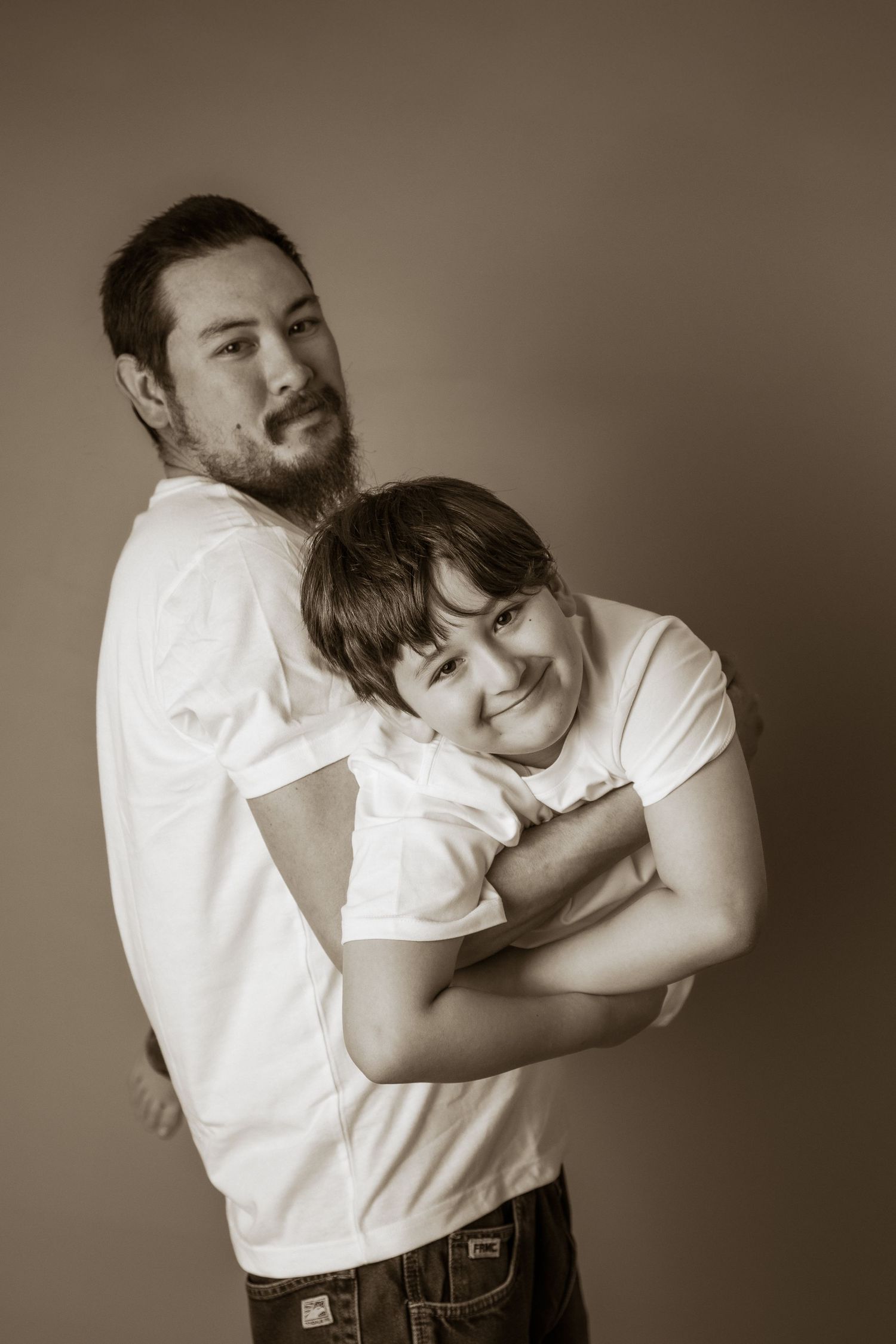 Sepia-toned portrait of a bearded man in white t-shirt hugging a smiling young boy, studio background.