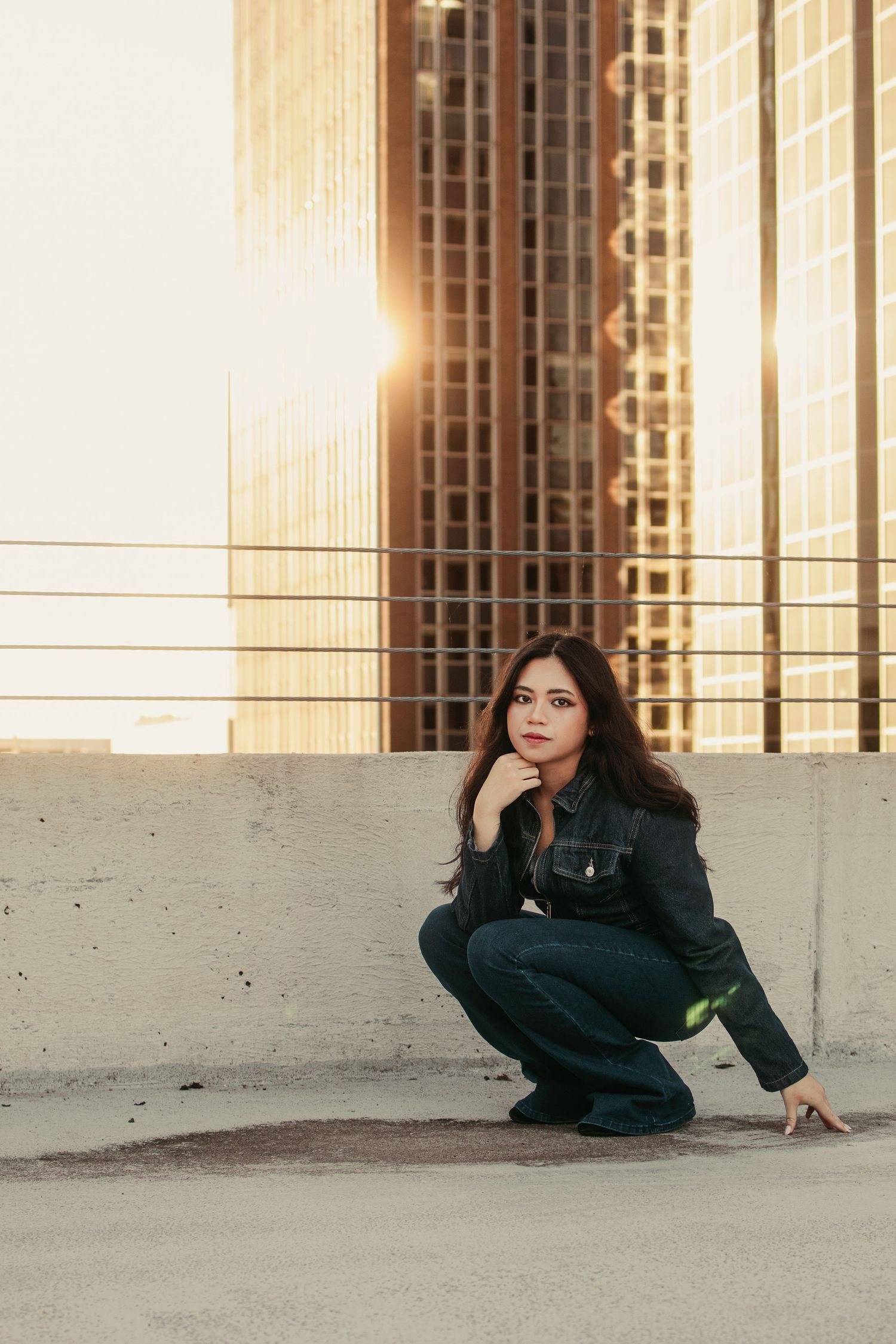 Young woman in dark denim crouches on rooftop at sunset with glowing skyscrapers in the background.