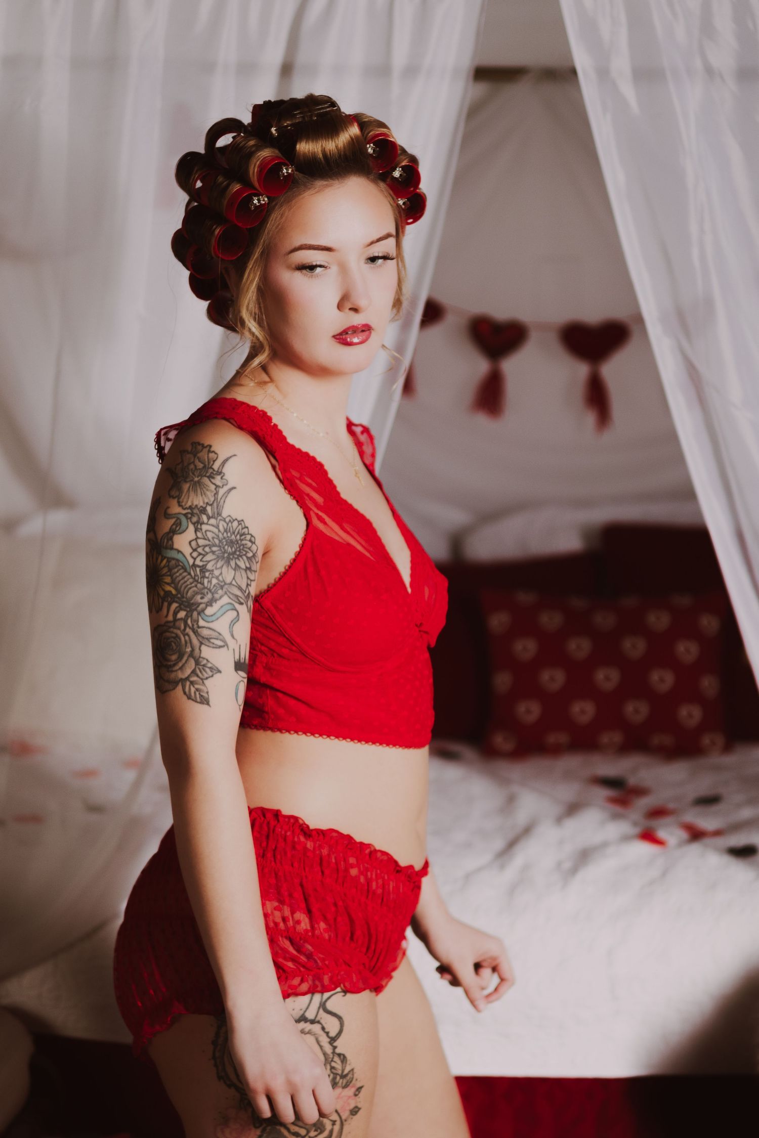 Woman in red lingerie set with hair curlers and tattoos poses in front of a canopy bed.