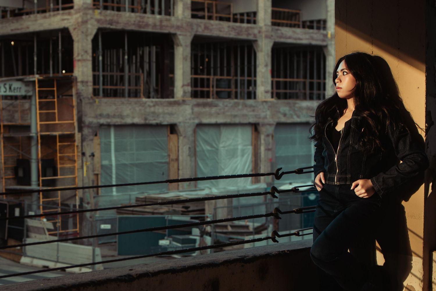 Woman in black jacket stands on urban rooftop gazing at abandoned building in warm sunlight.