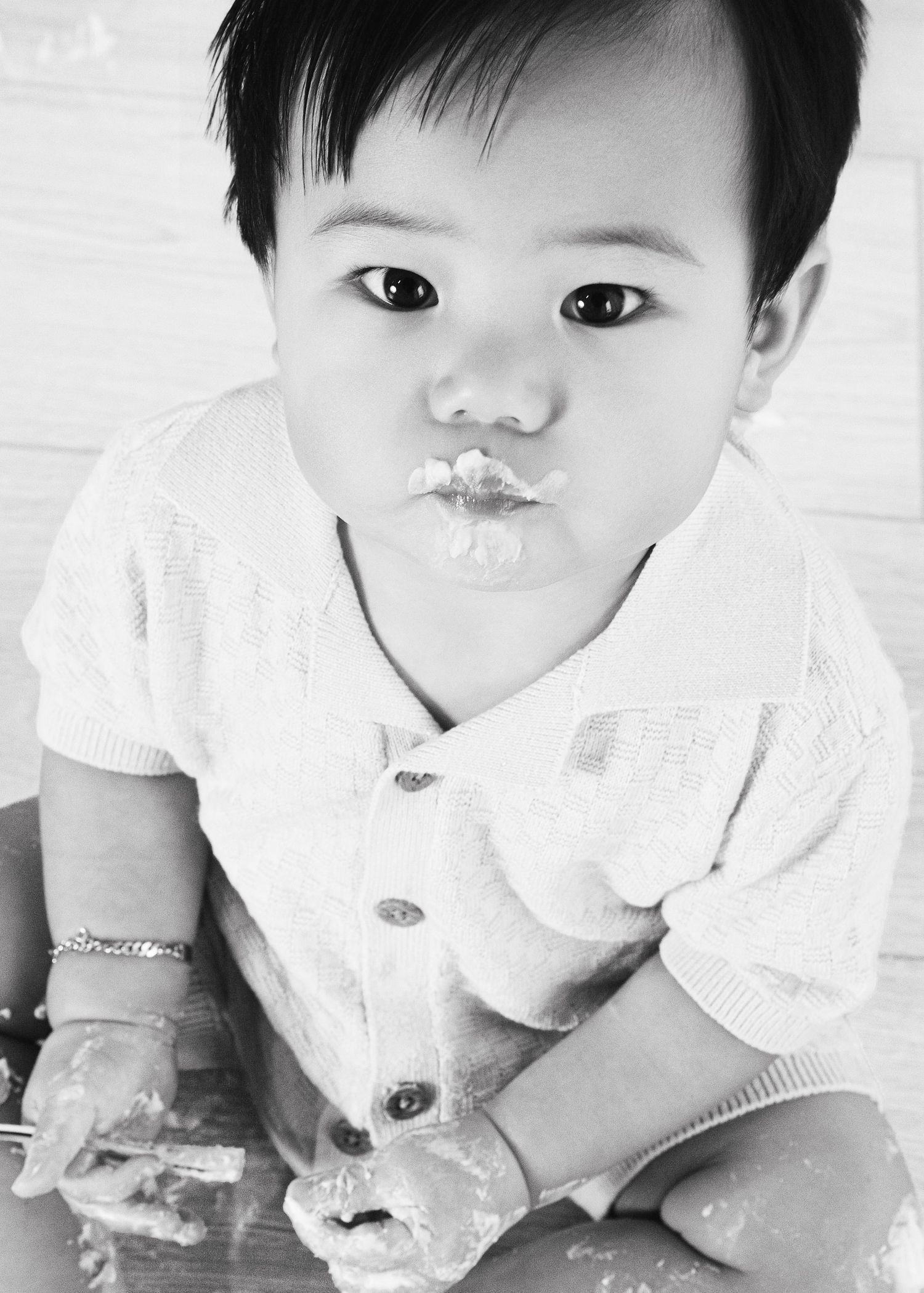 Black and white photo of a cute Asian baby with messy hands and frosting on lips, wearing a white knit cardigan.