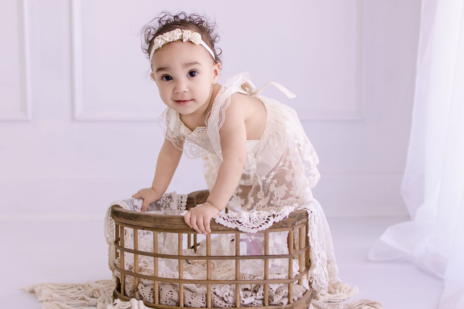 A cute baby girl in a white lace dress and headband stands in a rustic wooden basket in a bright studio.