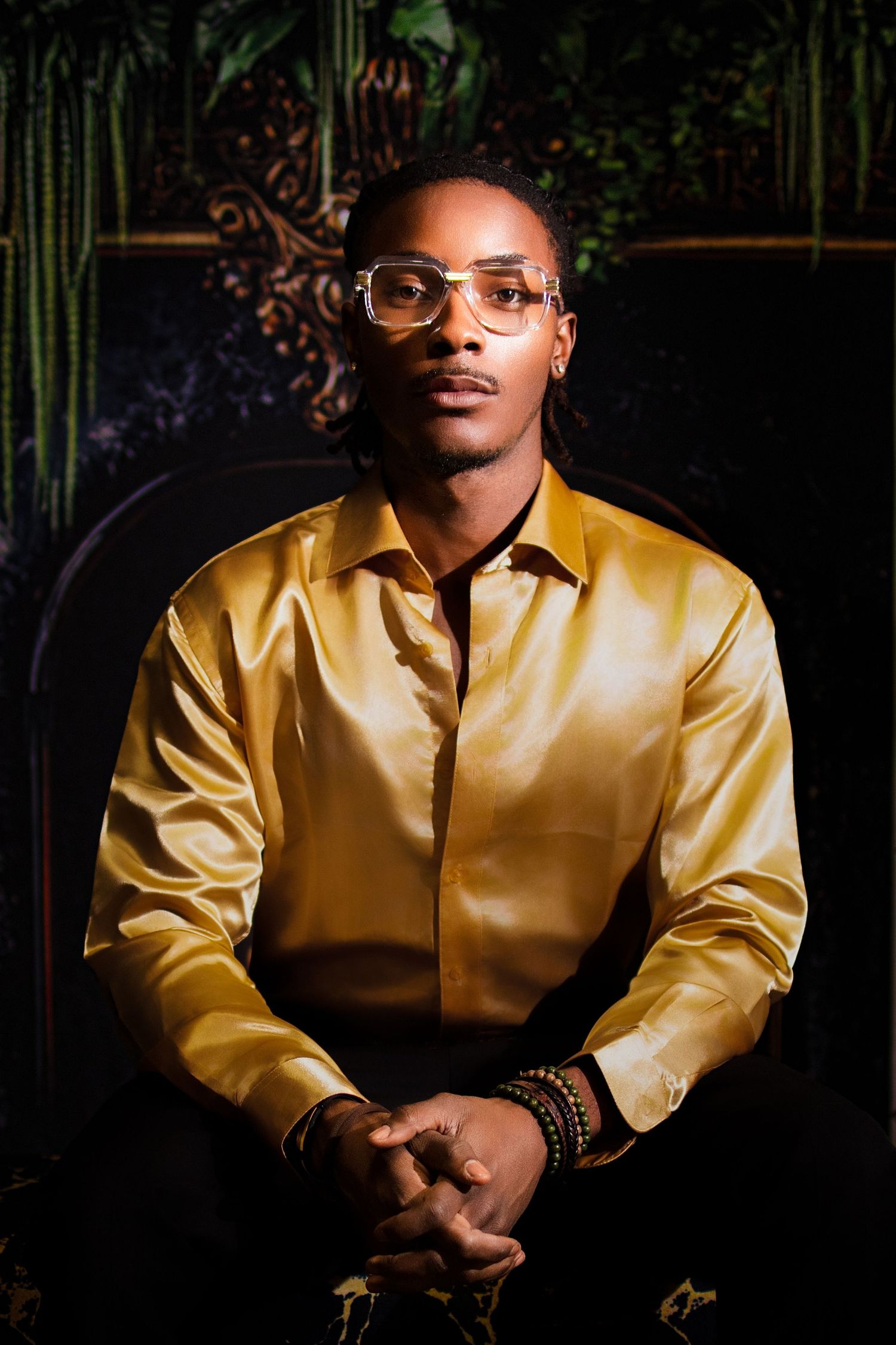 Stylish Black man in gold satin shirt and clear glasses sitting confidently in dramatic dark setting.
