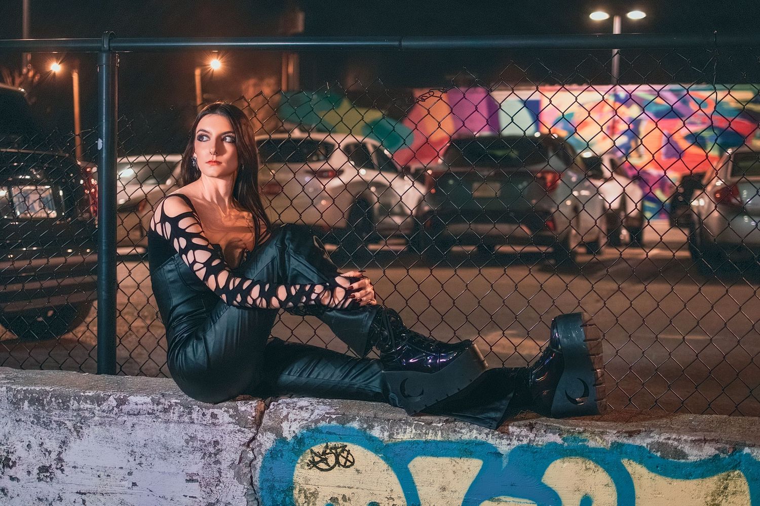 Woman in black leather outfit sitting against graffiti wall by chain-link fence at night.