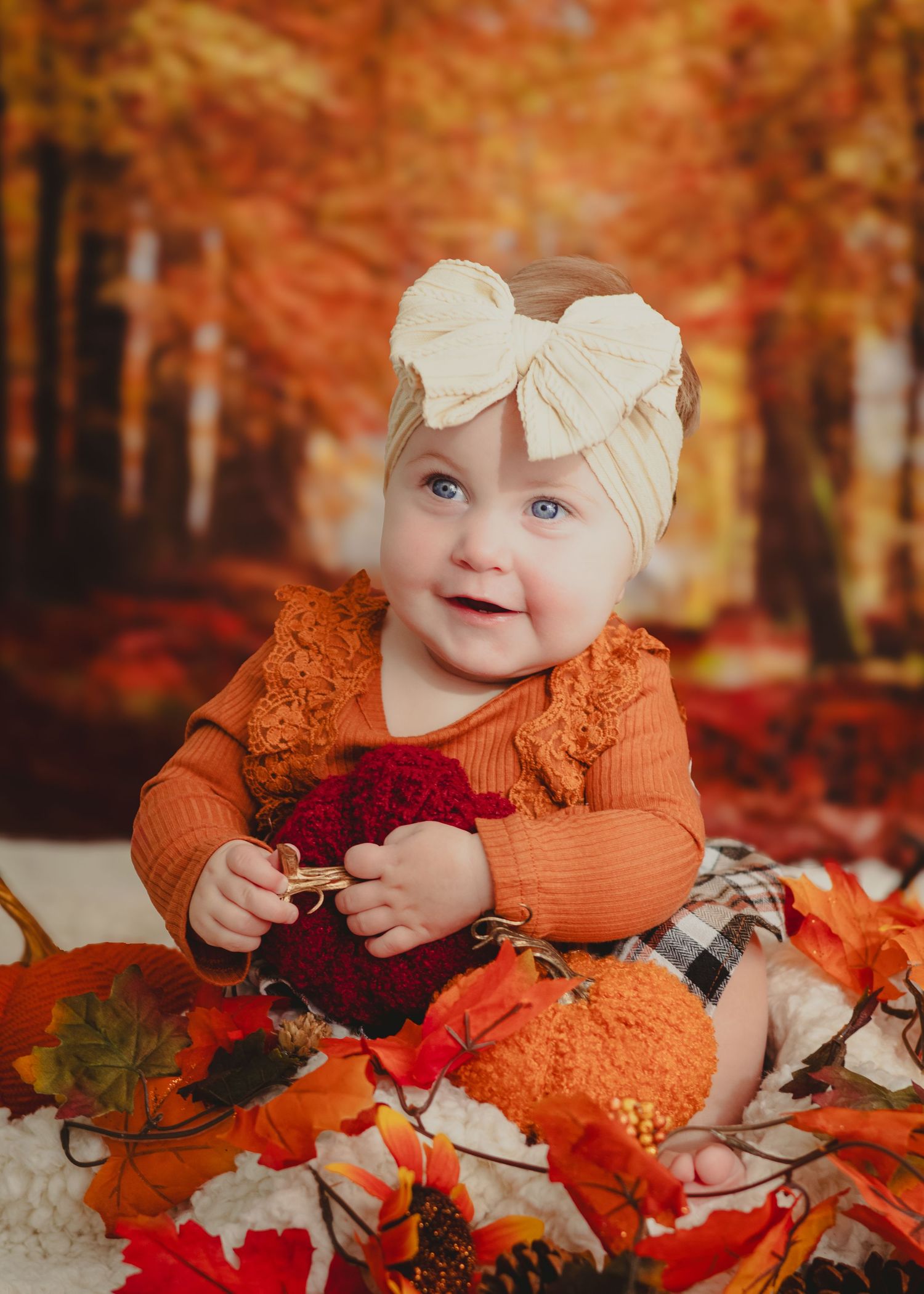 Smiling baby in orange outfit and cream bow headband surrounded by fall leaves with autumn forest backdrop.
