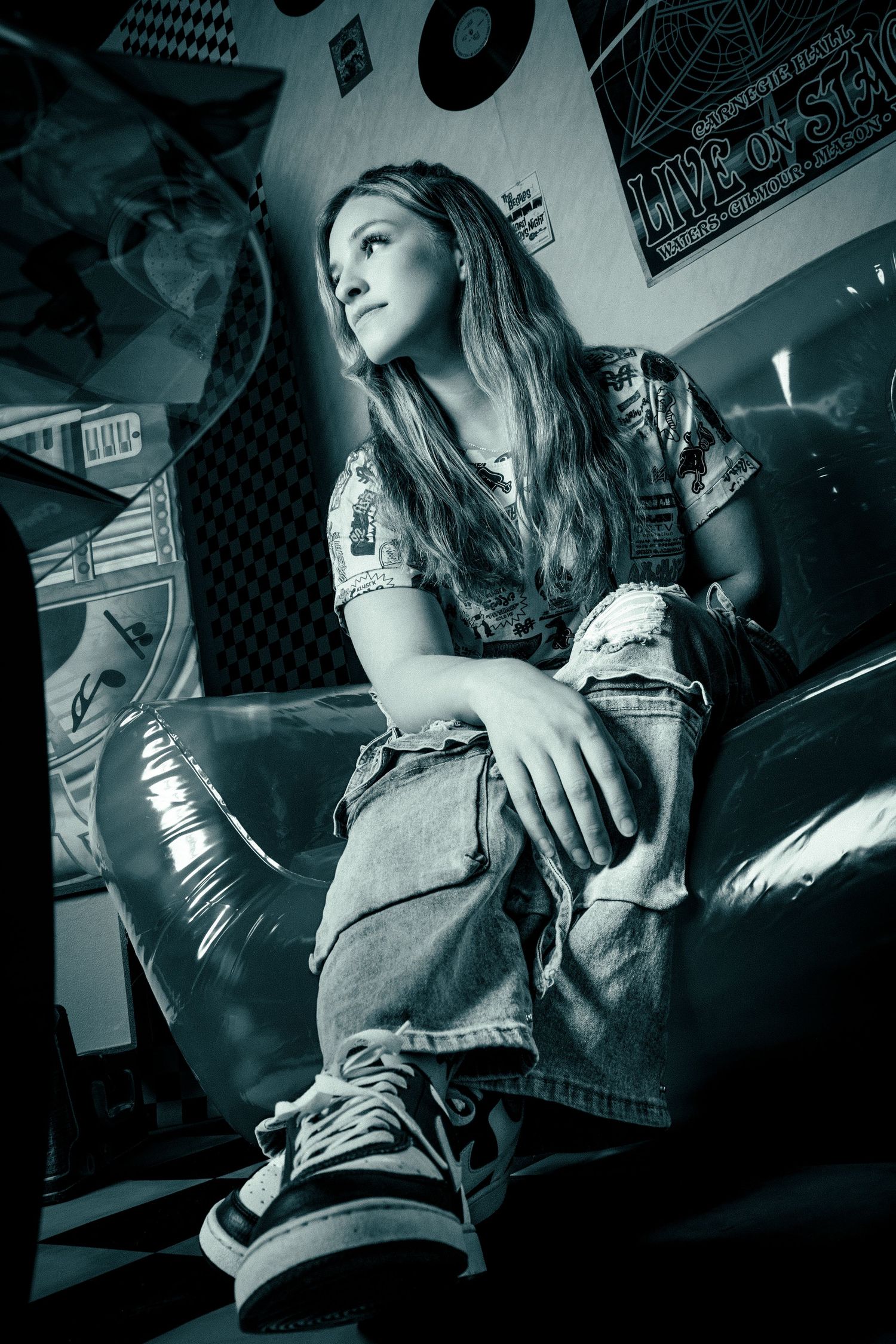 Black and white photo of teen girl in ripped jeans and sneakers sitting on pink inflatable sofa.