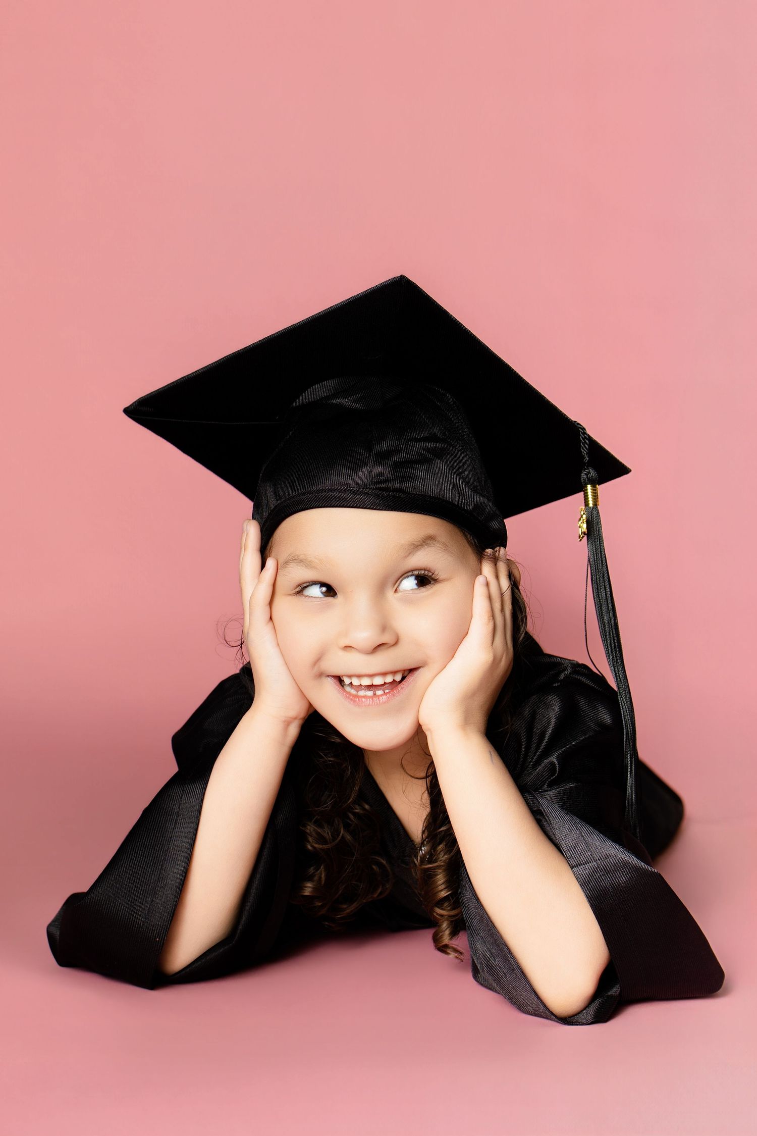 Happy young girl in black graduation cap and gown lying down with hands on cheeks on pink background.